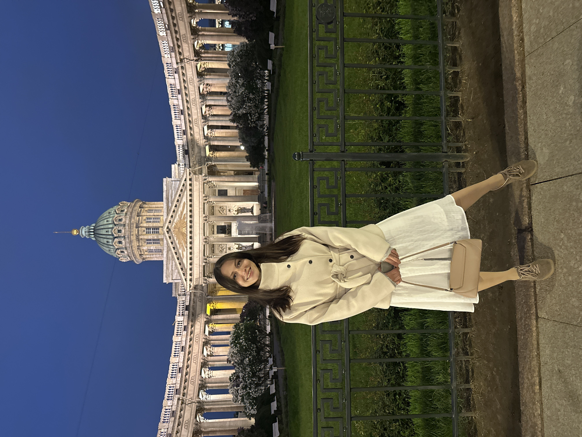 A long shot, from a low angle, shows a woman standing in front of the Kazan Cathedral, Saint Petersburg, Russia. The woman is positioned slightly off-center to the right, while the cathedral occupies the left half of the image. The cathedral's green dome is visible against a blue sky. The woman is wearing a white dress, a beige coat, and tan shoes, and she is holding a beige purse. A black metal fence runs behind her. Green grass is visible behind the fence, along with trees and benches. The image is taken at night, and the cathedral is illuminated, revealing the architectural details of its columns.