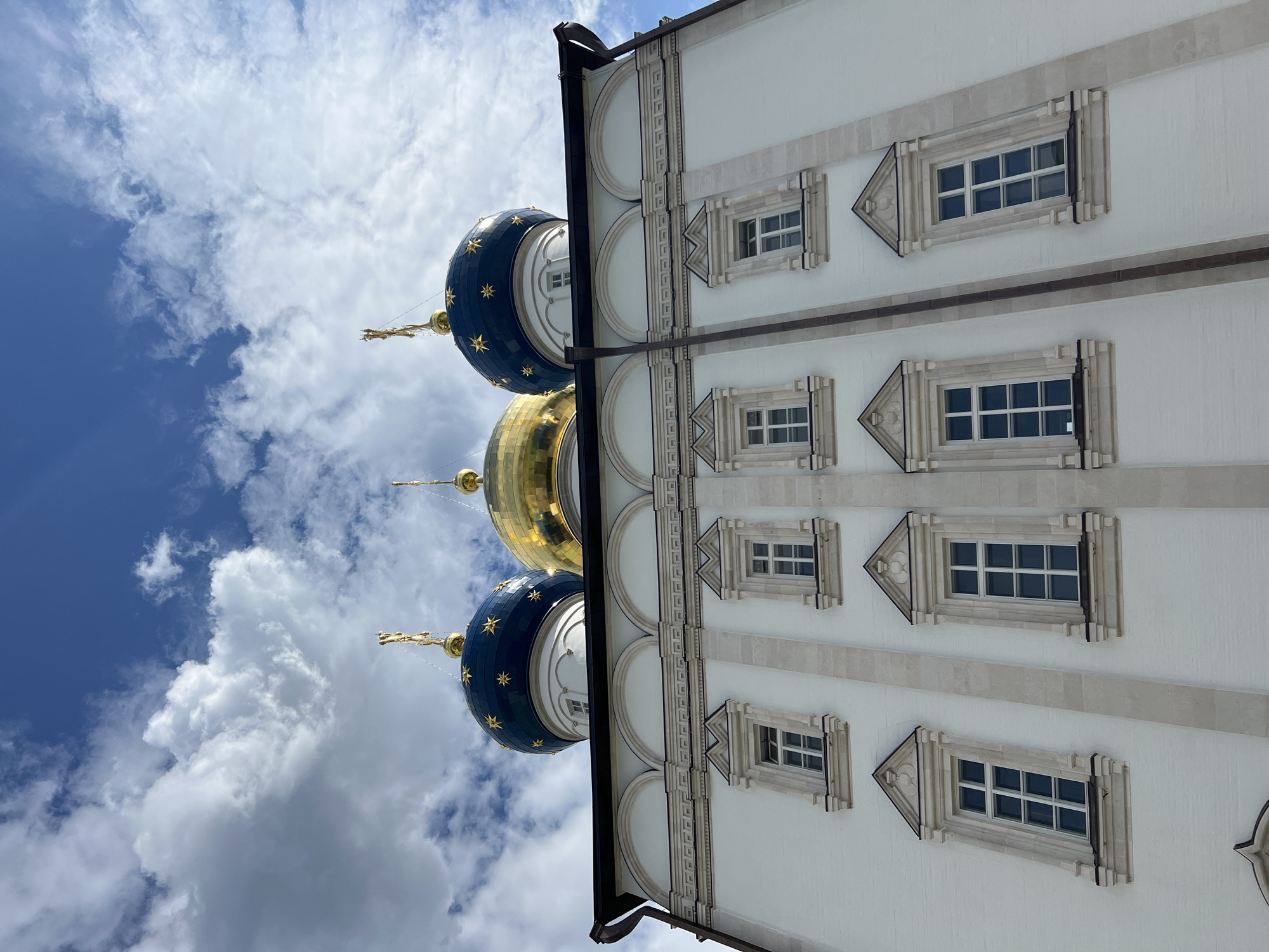 This is a low-angle, full-shot image of the Assumption Cathedral in Tver, Russia, captured on a partly cloudy day. The cathedral's white facade dominates the lower half of the frame, with multiple rows of rectangular windows. These windows are adorned with light stone frames, each topped with a small triangular pediment. The upper portion of the building features arched recesses, above which are three domes. The central dome is golden, while the two flanking domes are dark blue and decorated with gold stars. All domes are topped with golden crosses. The sky above is a vibrant blue, with scattered white clouds. The image appears to have been taken from ground level, looking upwards towards the cathedral.