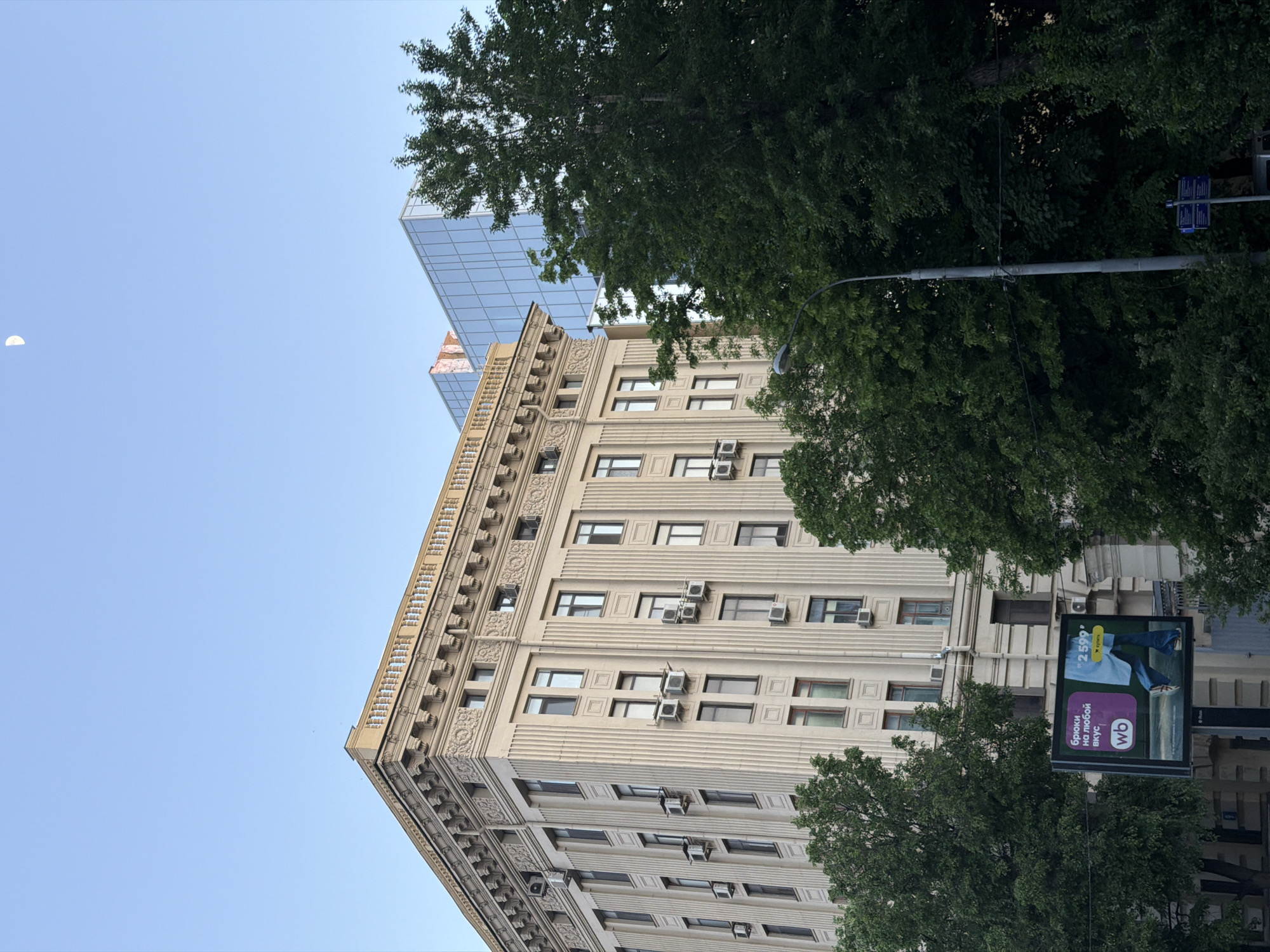 Here is a forensic description of the image:
The image shows a building in Moscow, Russia, viewed from a low angle. The building is light in color with multiple rows of windows and is partially obscured by trees. It is topped by an additional glass structure. A street lamp and advertisement for "WB" (likely Wildberries, a Russian online retailer) are visible. In the sky, a crescent moon is seen.