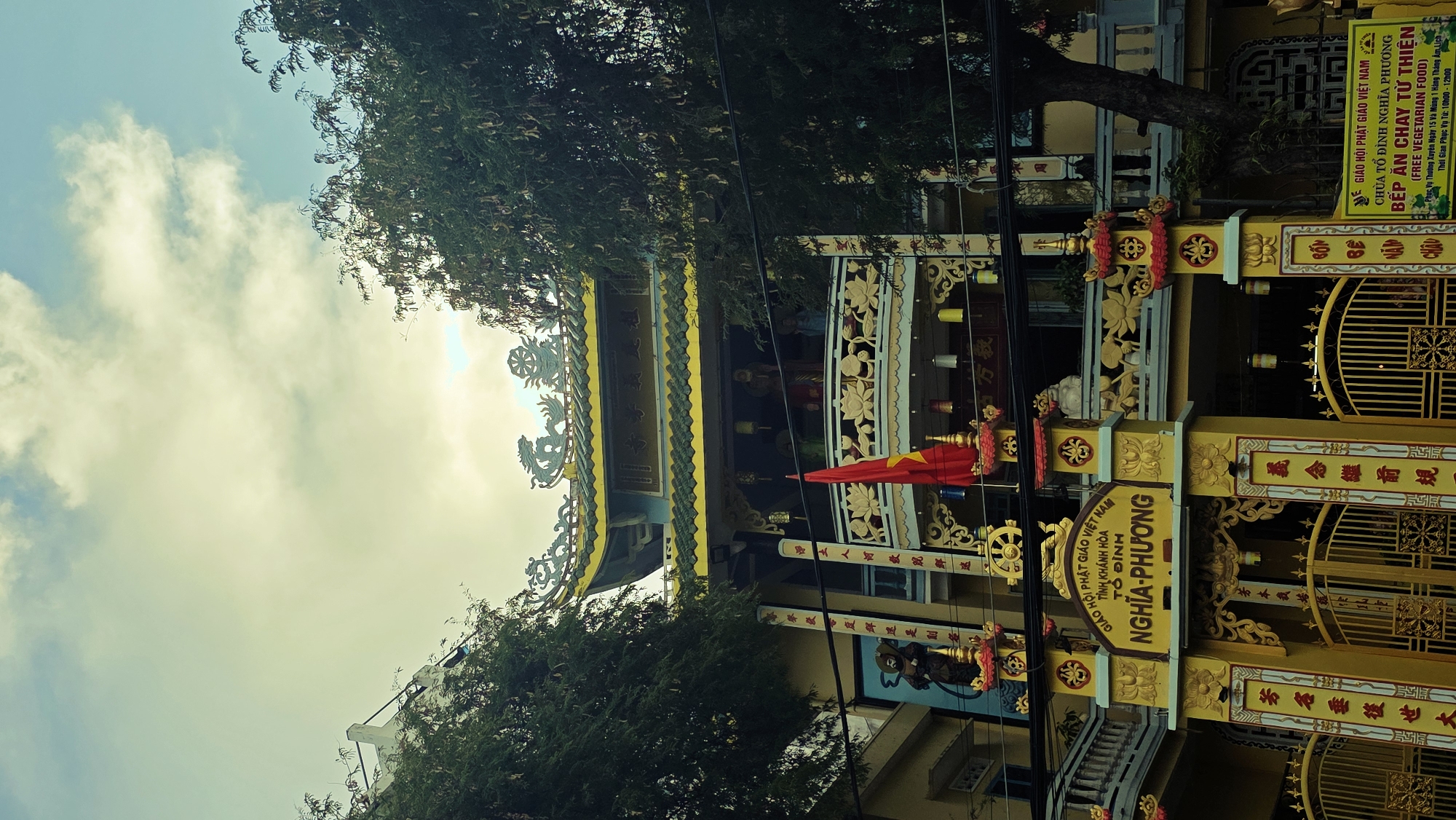 The image shows a building in Nha Trang Ward, Vietnam, likely a temple or religious site, based on the architectural style and signage. The building is painted predominantly in yellow and gold, with intricate decorative details. A Vietnamese flag is prominently displayed. Trees partially obscure the view of the building and the sky above is cloudy. Signage in Vietnamese script is visible. One sign identifies the location as "Tổ Đình Nghĩa Phương" (Nghĩa Phương Temple) and another indicates it offers free vegetarian food.