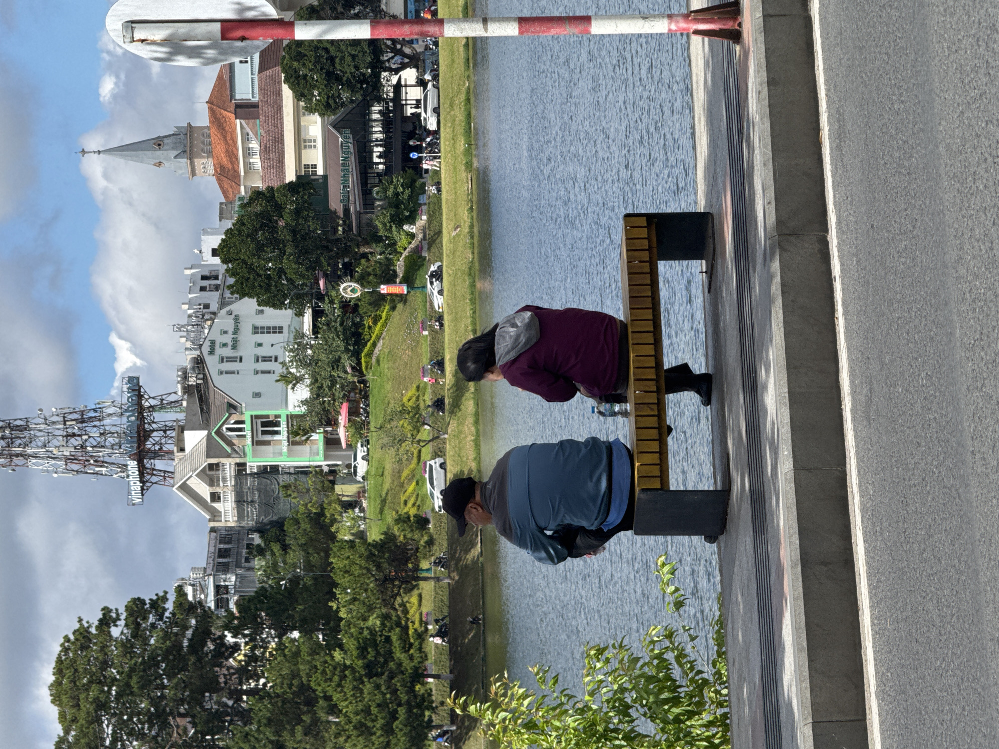 Two people are sitting on a wooden bench facing a lake. A man in a blue jacket and black baseball cap is seated on the left, and a woman in a burgundy jacket with a grey hood is seated on the right. The bench is situated on a stone platform or sidewalk, next to a paved road. Beyond the lake, there is a green grassy area with scattered trees and buildings, including a structure that resembles a church tower. In the background, a communications tower is visible, and buildings with various architectural styles can be seen. A red and white striped bar spans across the top of the image. The scene is in Da Lat, Vietnam, by Xuan Huong Lake.