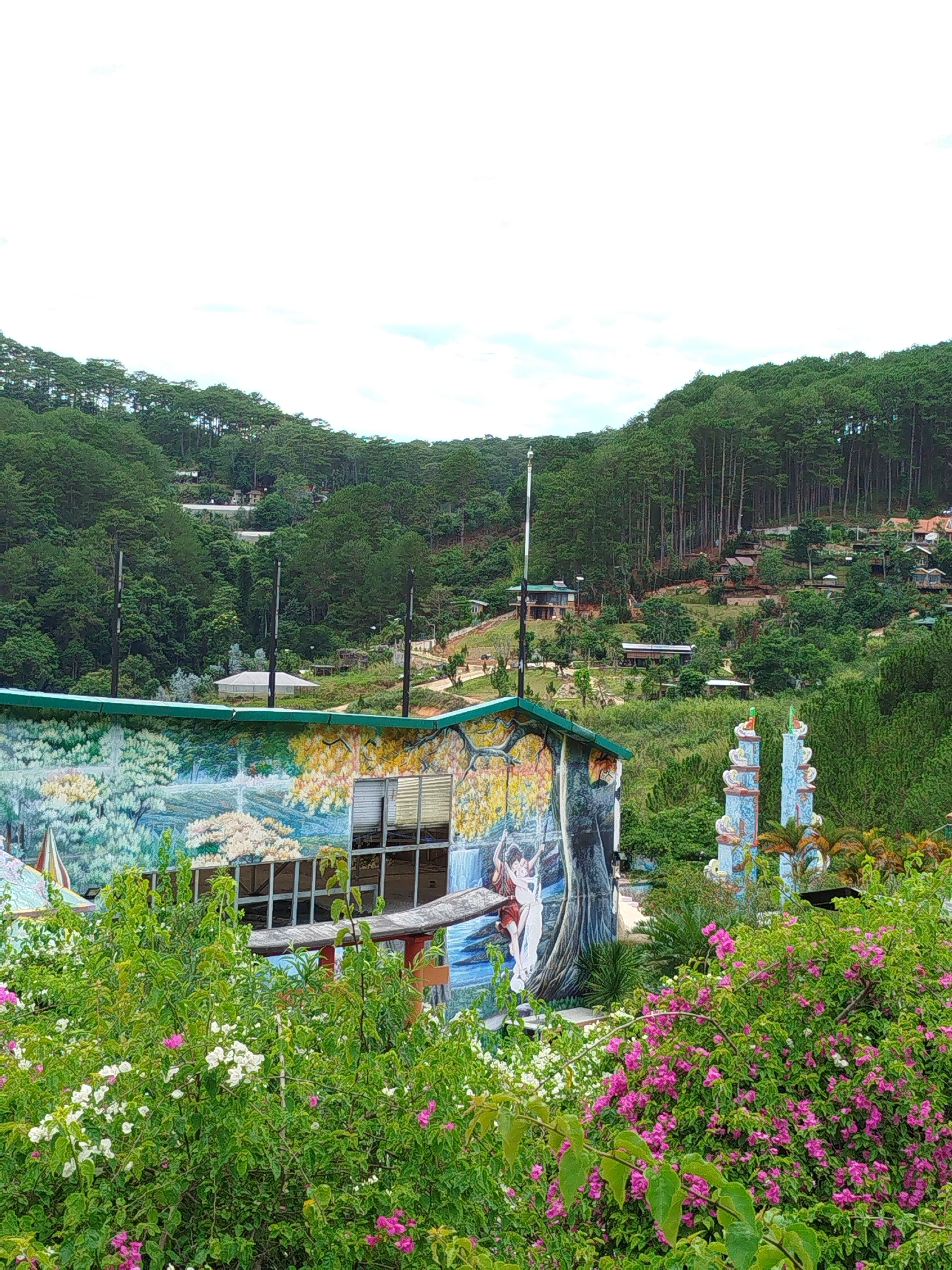 A medium, outdoor shot features a building with a landscape mural and two decorative columns in the foreground, against a backdrop of trees and other buildings. The image was taken in Da Lat, Vietnam. The building in the foreground has a green roof and a mural that includes trees, flowers, and figures. Two decorative blue and white tiled columns stand beside it. In front of the building, a hedge of flowering bushes with white and pink blossoms partly obscures the view. In the background, a dense, verdant hillside is dotted with homes, a pavilion, and trees of varying sizes. The sky is partly overcast.