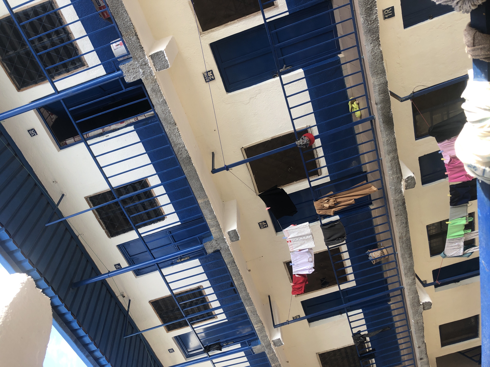 The image depicts the exterior of a multi-story building, likely an apartment complex or dormitory, with a distinctly tilted perspective suggesting it was taken from a low angle. The building's facade is painted white with a prominent blue trim along the balconies and window bars. Various rooms are visible, each marked with a room number, such as "3M," "3R," "2F," and "1R." Laundry is hung out to dry on lines suspended between balconies, with various garments including shirts, shorts, and possibly undergarments. Some of the doors are open, revealing glimpses into the interiors. Yellow clogs are visible on one of the balconies. The environment appears to be a residential area within the city of Jos, Nigeria, characterized by the architectural style and the visible laundry, common in such urban settings. The sky above is a light blue with some white clouds. The image's composition and the presence of laundry suggest an everyday scene of domestic life.