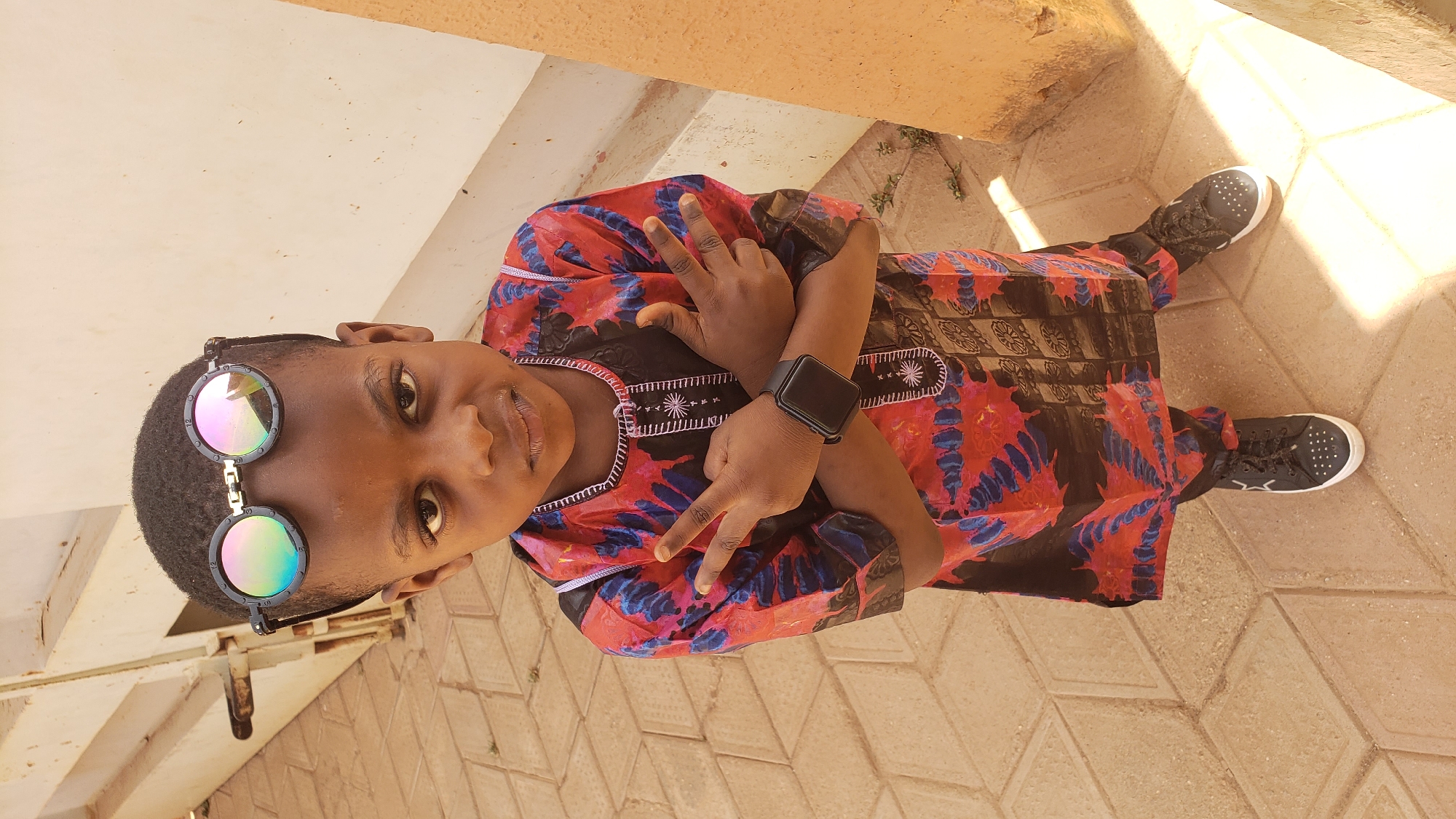 A young person stands outside in Sabon Tasha, Nigeria. They are wearing black and white sneakers, and an ornate patterned garment in red, black, and blue, with a watch on their left wrist. Sunglasses are perched atop their head. The person is making a hand gesture. The location appears to be an outdoor area with light-colored pavement and adjacent structures.