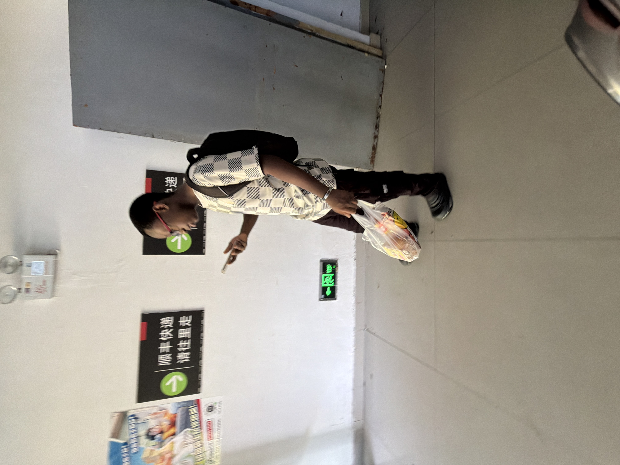 The image shows a young Black man in a hallway, standing with his side to the camera. He is wearing glasses, a checkered shirt, dark pants, and black shoes. He is carrying a plastic bag with food items in his left hand and holding a smartphone in his right hand, appearing to be looking at it. A black backpack is on his shoulders. Behind him, on a white wall, are two black signs with white Chinese characters and arrows, indicating directions. One sign appears to be related to a delivery service. To the left of the signs is a poster or advertisement. A green illuminated exit sign is visible lower on the wall. The floor is tiled with light-colored tiles. To the right of the man is a large, grey, textured door or panel. The overall environment appears to be an indoor public space, possibly a transportation hub or commercial building. The context provided by the user indicates this location is the city of Jiangjin, China.