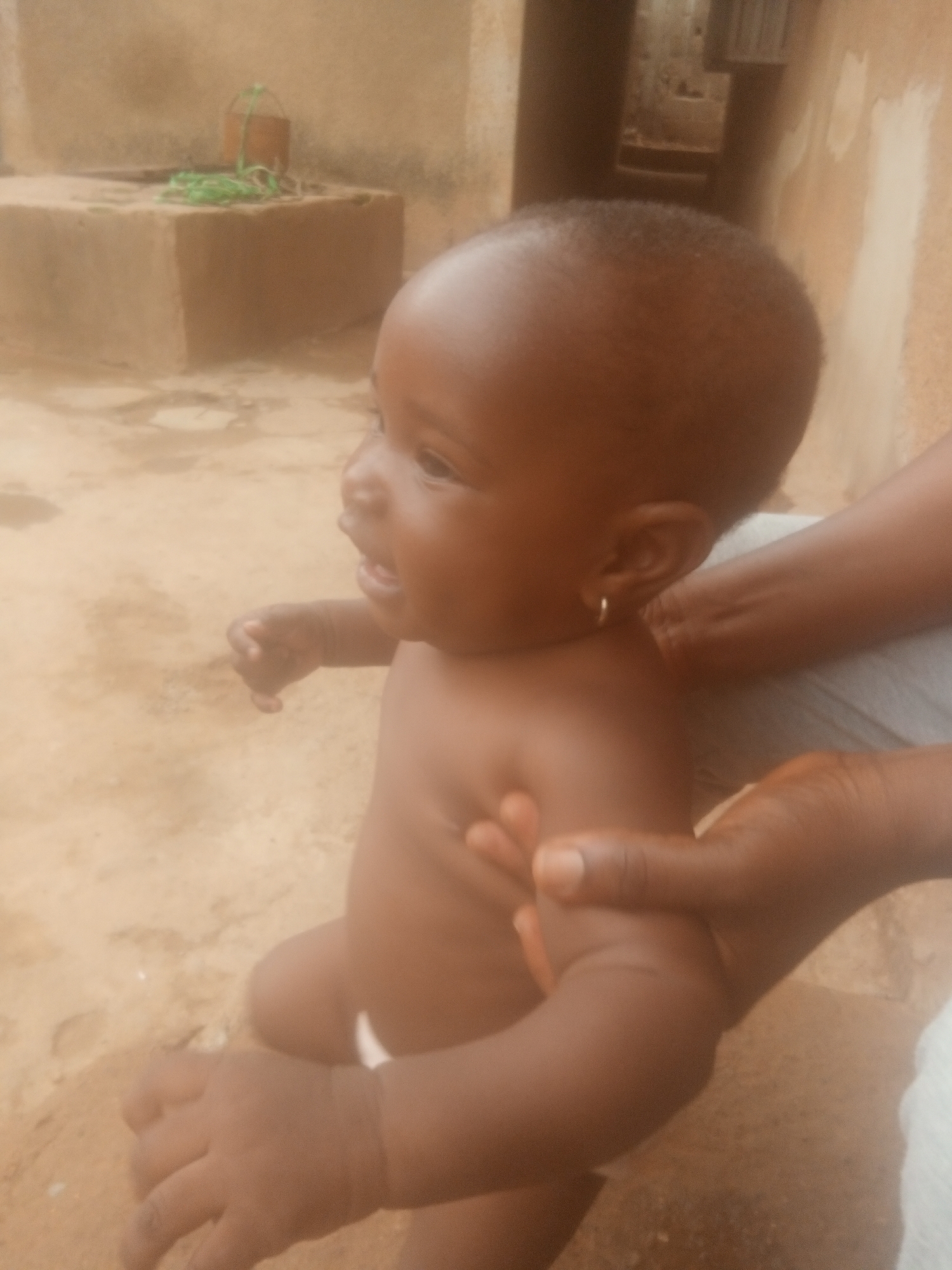 A baby sits on the lap of an adult in an outdoor environment. The baby has dark skin, short hair, and a gold earring in its left ear. The baby's hands are visible, with the fingers slightly curled. The adult's hands hold the baby's arms. The adult wears a light-colored garment. In the background, there is a concrete structure with green rope and a bucket on top. The location is in the city of Unknown, Unknown.