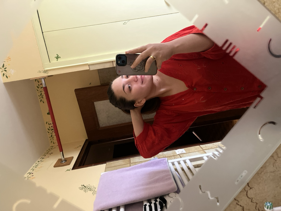 The scene depicts a mirror selfie taken in a domestic bathroom located in Ancenis, France. A female with medium-length dark hair, pulled back, is visible in the reflection. She is wearing a red, collared, long-sleeved blouse featuring a button-front closure and eyelet embroidery. Her left hand is positioned near the back of her head, while her right hand holds a dark-cased smartphone, used to capture the image. A gold-colored ring is present on her left ring finger. The mirror has an irregular, decorative shape with frosted or etched cut-outs along its lower and left edges. Reflected in the mirror, the bathroom walls are painted pale yellow with green and beige floral stencil patterns. A white ladder-style towel rack, holding a light purple towel and a black-and-white striped towel, is visible on the left. Above it, a red-covered metal bar, secured by a wooden mount, extends into the corner. A dark wooden door frame is partially visible in the background. To the right, a light-colored two-door cabinet, also adorned with small green stencil designs, is reflected.