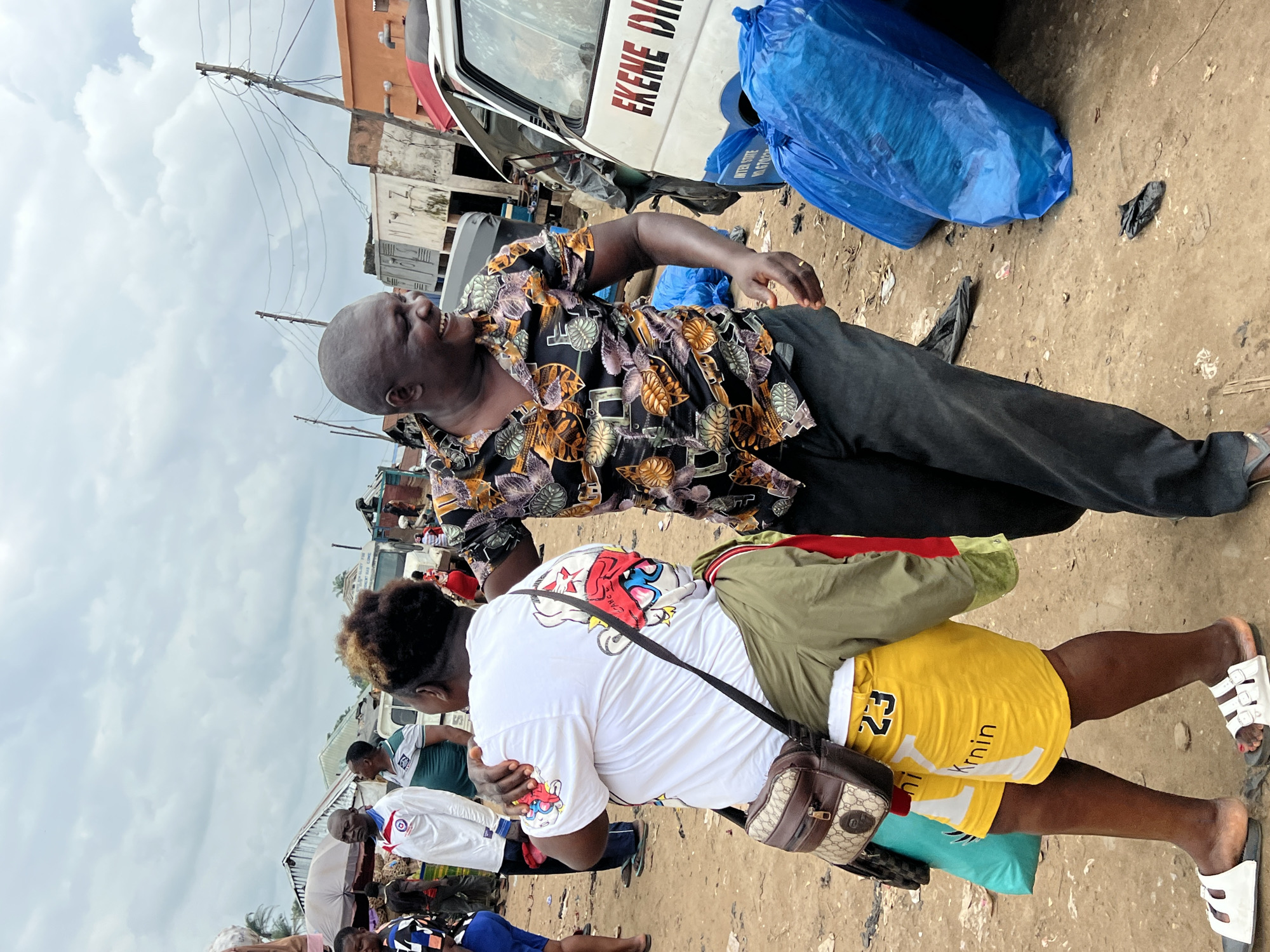 In Oko Amakom, Nigeria, a man and a woman are interacting in a market. The man, with a shaved head, is wearing a black and gold patterned short-sleeved shirt and dark pants. He is laughing heartily, with his right arm extended towards the woman. The woman, with dark, styled hair, is wearing a white t-shirt with a cartoon graphic and yellow shorts with the number "23" on them. She has a brown and beige patterned shoulder bag. They appear to be in a lively exchange, possibly amidst the bustle of the market. The ground is a dirt surface, littered with some debris. In the background, a white van with "EKENE DIR" written on its side is visible, along with various market stalls and structures. Several large blue plastic bags are also present. The sky is overcast with clouds.
