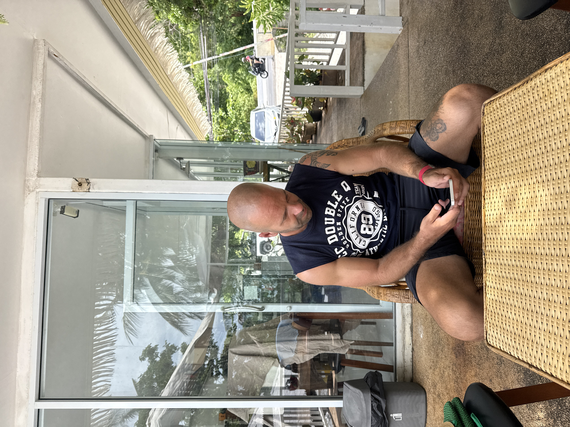 A bald, fair-skinned man with a muscular build is sitting in a wicker chair on a covered patio in Ko Samui District, Thailand, looking down at a smartphone in his hands. He is wearing a dark blue sleeveless t-shirt with white lettering and graphics, and dark blue shorts. A bright pink wristband is visible on his left wrist. The patio has a woven rattan table in front of the man, and a trash can is to his left. Behind the man, large glass doors reflect palm trees and the sky, suggesting an outdoor or semi-outdoor establishment. Further in the background, a white car and a person on a motorcycle are visible on a road lined with lush green foliage. The overall environment appears to be warm and tropical.