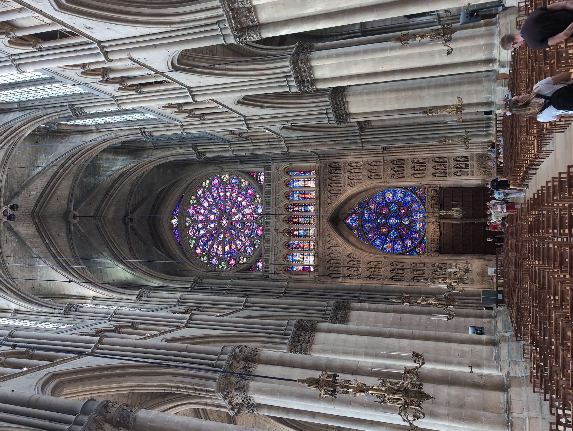 This is an interior shot of a large Gothic cathedral, specifically the Reims Cathedral, located in the city of Reims, France, not Urvillers. The perspective is from the nave, looking towards the altar and the stained-glass rose windows. Massive stone columns with intricate carvings line both sides of the nave, supporting ribbed vaulted ceilings. Two large, ornate stained-glass rose windows, one above the other, dominate the far wall, displaying a vibrant array of colors. Below the upper rose window are rectangular stained-glass panels. Rows of empty wooden chairs are set up in the nave, with a few people walking in the background near the altar area. The overall impression is one of grandeur and historical significance.