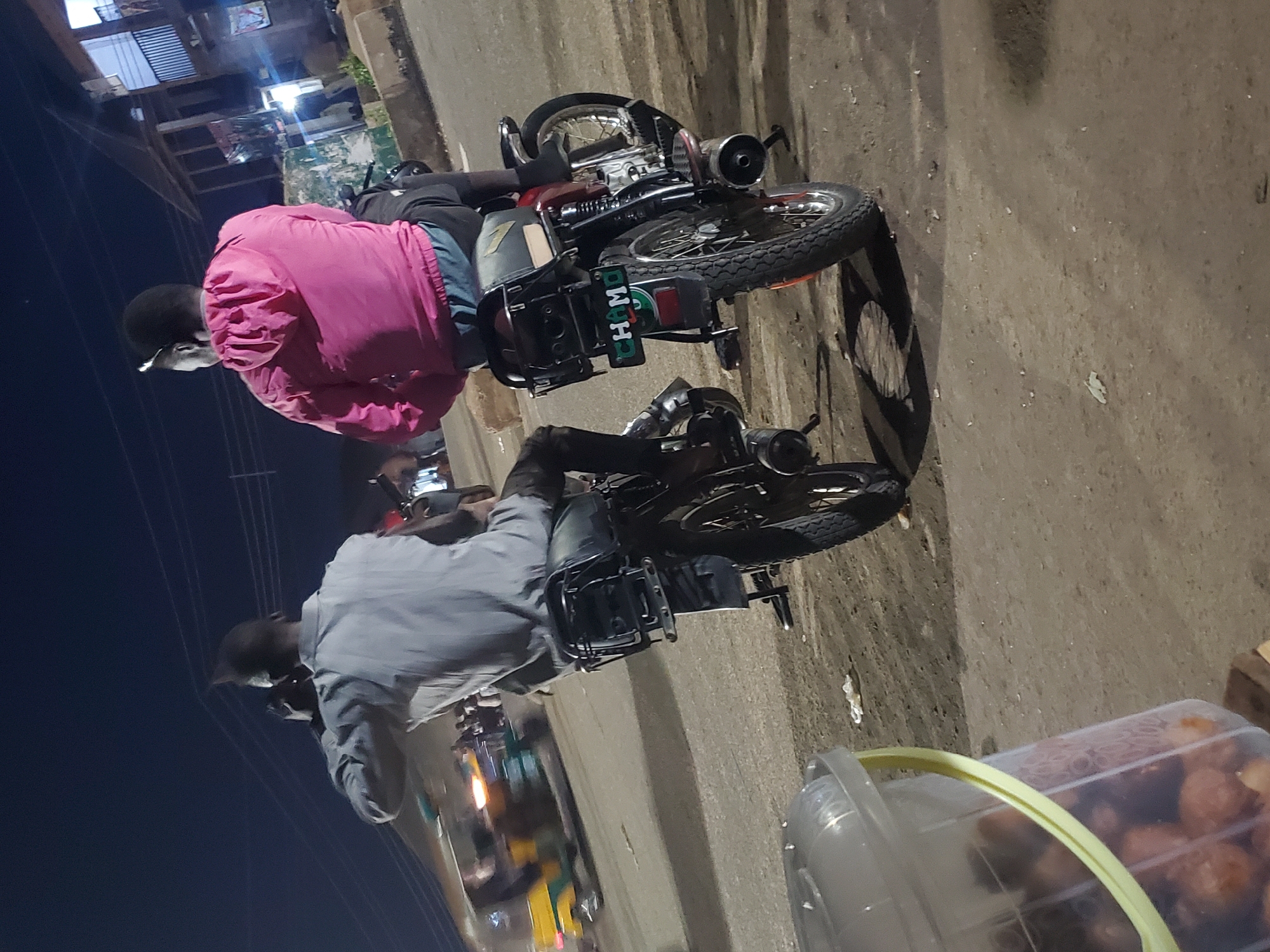 Two individuals are seated on separate motorcycles, facing away from the camera, at night. The person on the left is wearing a light blue collared shirt, and the person on the right is wearing a pink hooded jacket. Both individuals appear to be engaged in conversation or observing something. The motorcycles are positioned side-by-side on a dirt and asphalt road. In the background, there are buildings, streetlights, and a blurred yellow and green vehicle, possibly a tricycle, suggesting an urban or semi-urban environment. A clear plastic bucket filled with what appears to be fruit is in the foreground to the left of the frame. The overall scene depicts a nocturnal street setting in Gwong, Nigeria.