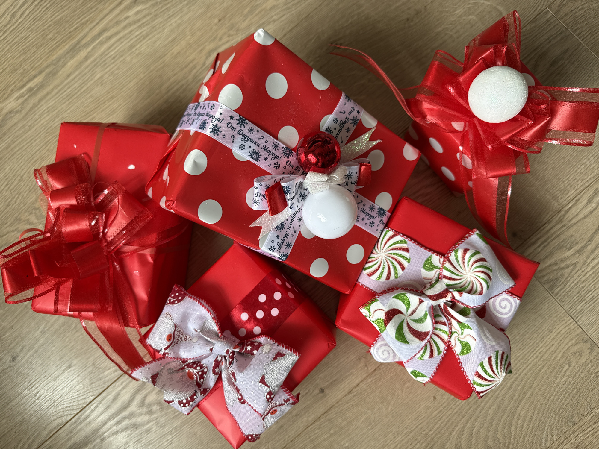 The image shows a collection of five Christmas presents on a light-colored wooden surface. The presents are all wrapped in red paper with varying patterns: one has white polka dots, two are plain red, and two have a red and white polka dot panel. Each present is adorned with a decorative bow made of different ribbons. One bow is red with a white styrofoam ball in the center. Another is a combination of red, white, and green ribbons resembling peppermint candies. The other two bows are white with red and silver accents. One present is wrapped with a ribbon that reads "От Дедушки Мороза!" (From Grandfather Frost!). There are no indications of the scene's location in Votkinsk, Russia.