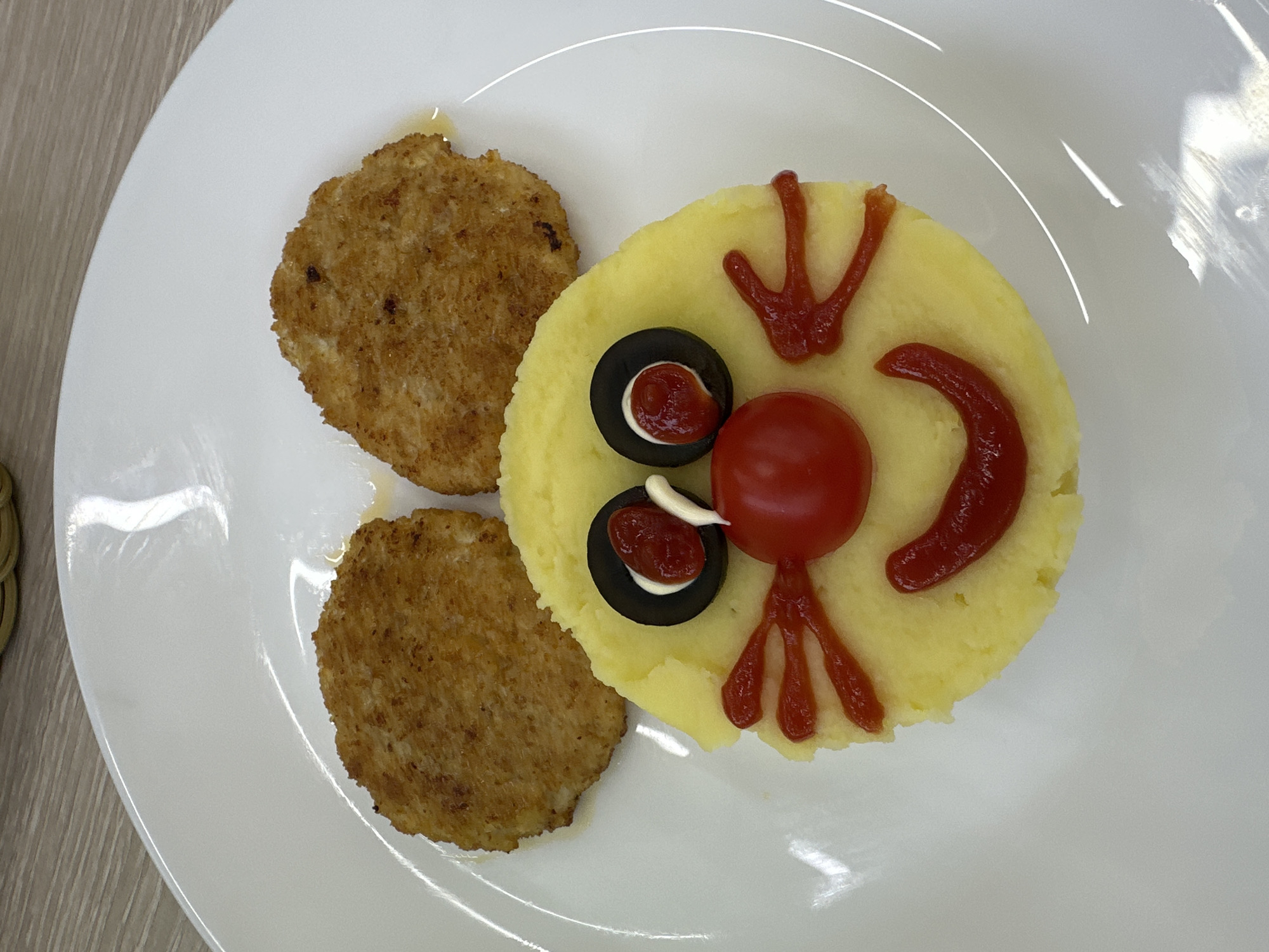 This media file shows a close-up, overhead shot of a white plate containing a meal creatively arranged to resemble a face. The main subject is a circular mound of mashed potatoes, serving as the "face." Two golden-brown, circular patties, likely meat or vegetable, are placed to the left of the mashed potatoes.
The mashed potato "face" is decorated with various food items to create features. For the eyes, two slices of black olives are used, each with a dollop of white sauce and a small amount of red sauce inside. A cherry tomato is positioned in the center, likely representing the nose. Red sauce is artfully applied to form a "mouth" in a crescent shape to the right of the tomato and "legs" or "feet" extending downwards from the tomato. The overall presentation suggests a playful and child-friendly meal.
The setting appears to be a table, with a wooden surface visible in the background on the left. The lighting suggests indoor illumination, possibly during the day. No text is visible in the image. The meal is the sole focus, arranged on a plain white plate with subtle curved indentations. The context of Chaikovsky, Russia, is provided.
