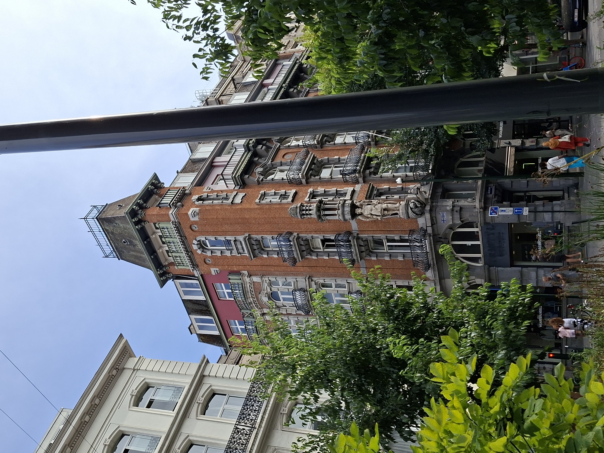The image shows a street scene in Brussels, Belgium. Two historic buildings with ornate architecture, one a light-colored facade and the other a red brick facade with a prominent turret, are visible. Trees with lush green foliage partially obscure the view of the buildings. In the foreground, a dark vertical pole dominates the right side of the frame. Several people are walking on the sidewalk in front of the buildings, and there are shop windows visible at street level. One shop has signage that reads "WEAREBRUXELL." The sky is a clear blue with a few faint horizontal lines that could be wires. The overall impression is a sunny day in an urban environment with a mix of architectural styles and pedestrian activity.
