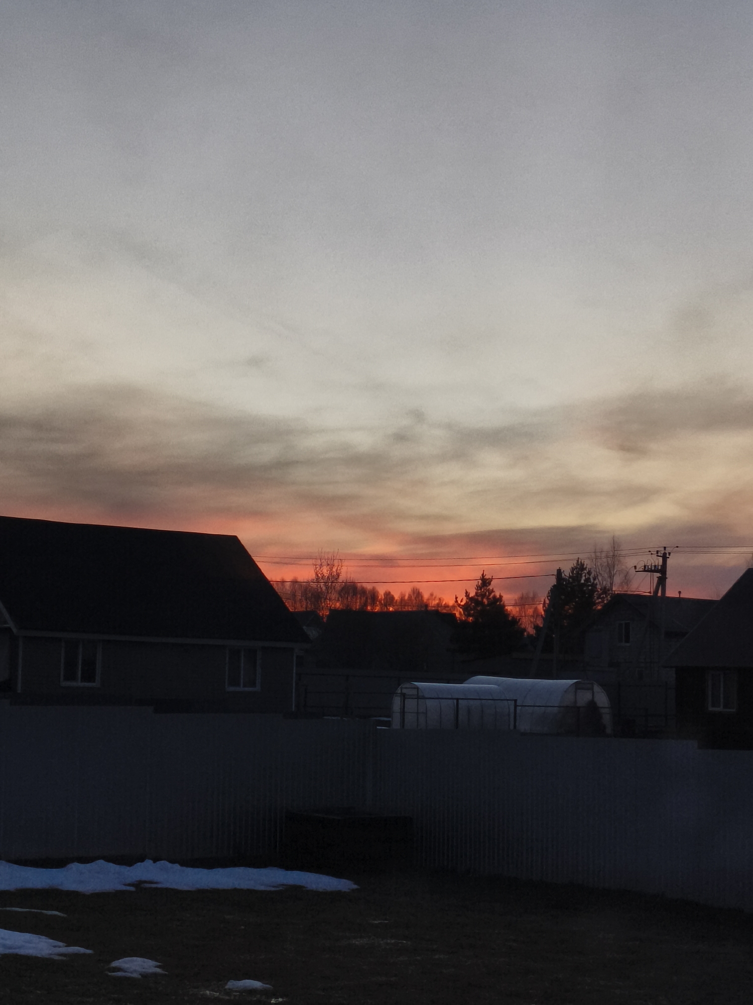 The image captures a twilight or sunset scene in Русский Вожой, Russia, with the sky exhibiting a gradient from pale grey at the top to a vibrant orange-red on the horizon. A silhouette of treetops stands against the bright horizon. In the foreground, the dark outlines of houses are visible, along with a greenhouse, and a corrugated metal fence. Patches of snow linger on the dark ground. Electrical lines stretch across the horizon.