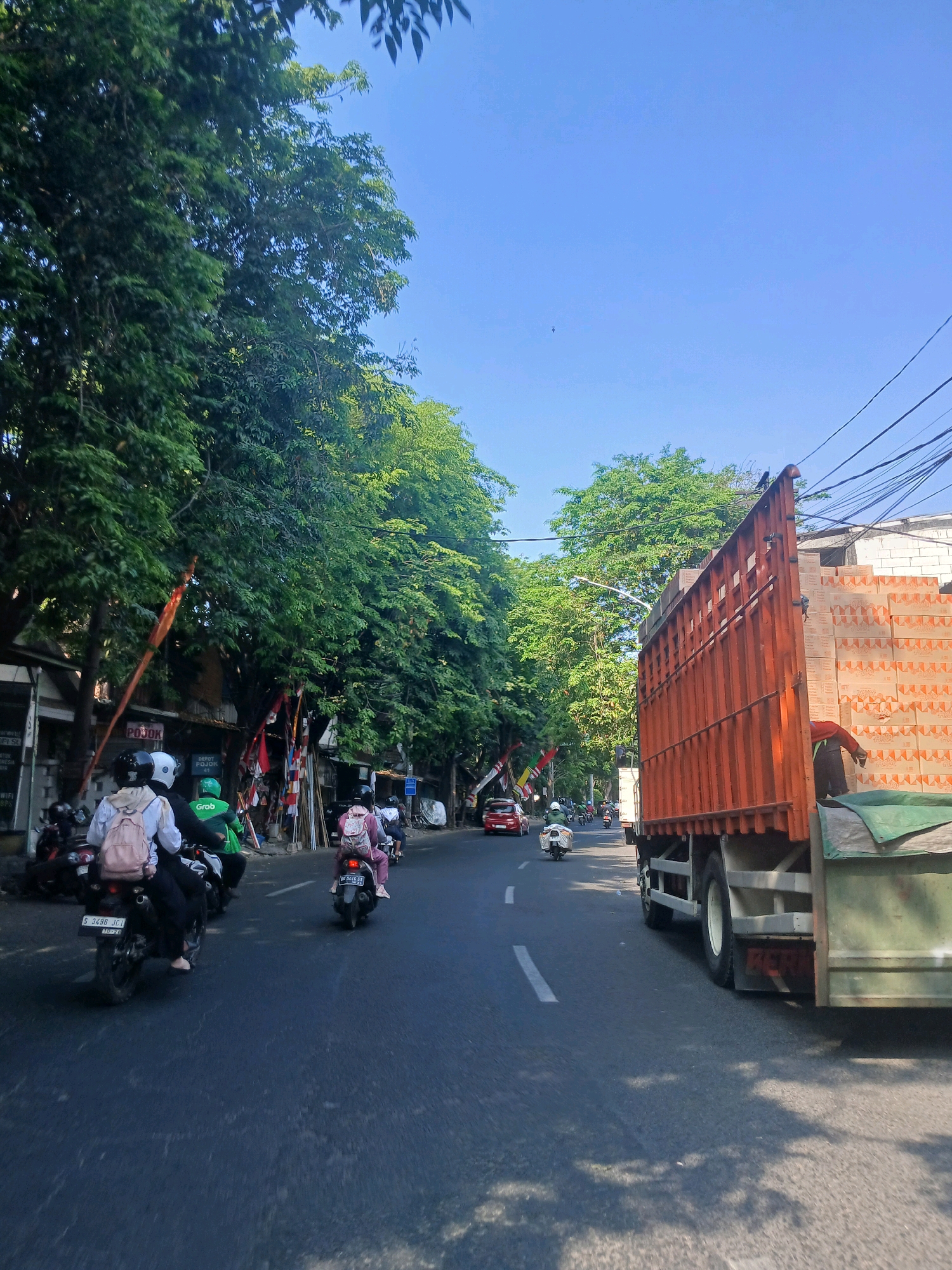 The image depicts a street scene in Surabaya, Indonesia, on a bright, sunny day with a clear blue sky. The perspective is from a moving vehicle, looking forward along an asphalt road lined with lush green trees on the left and a large orange truck carrying stacks of boxes on the right. Several motorcycles are on the road, with riders wearing helmets. On the left side of the road, there are buildings, more trees, and what appear to be flags or banners. A red car is visible further down the road, also heading in the same direction. The truck on the right is partially blocking the view of the road ahead. Electrical wires are strung overhead.