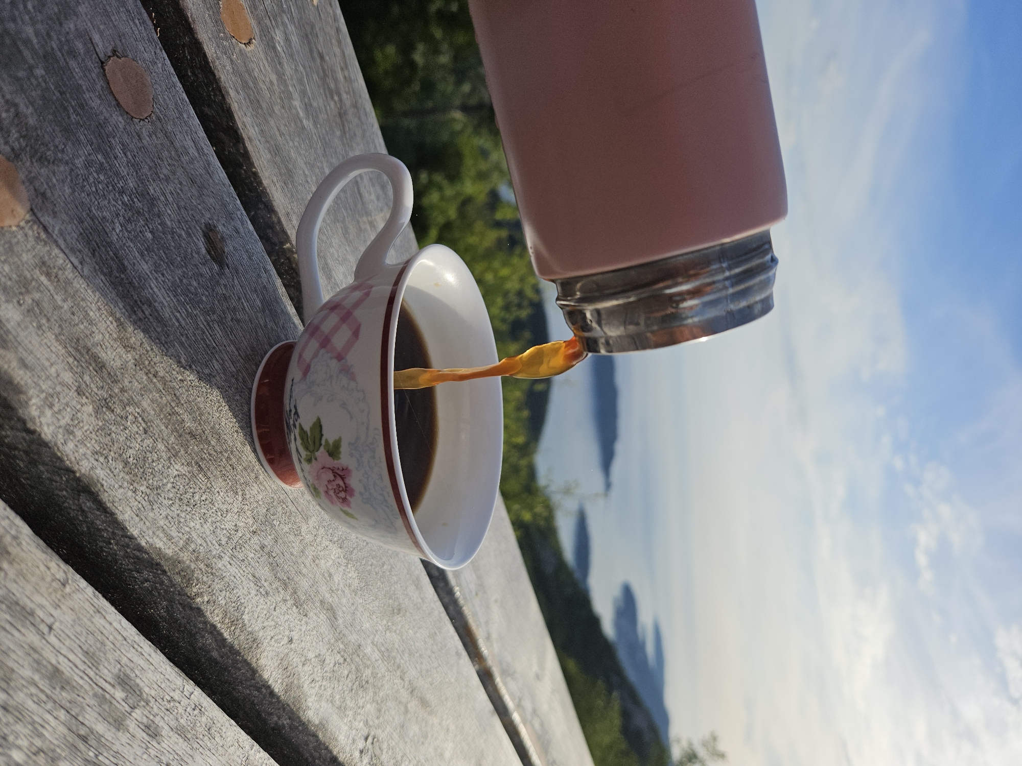 A pink insulated bottle with a silver rim is angled, pouring a stream of dark brown liquid, identified as coffee, into a white ceramic teacup. The cup features a delicate handle and is decorated with a floral pattern of pink roses and green leaves on its lower half, transitioning to a red grid pattern on its upper rim. The cup rests on a weathered wooden surface, possibly a table or bench, exhibiting prominent grain and a few flat, light-brown elliptical shapes scattered upon it. Direct sunlight creates distinct shadows on the wooden surface. The background reveals a sweeping natural landscape from a high vantage point, with lush green foliage covering a hillside that slopes down to a large body of calm, light blue water. Multiple landmasses, appearing as islands or peninsulas, protrude into the water, creating an intricate coastline. Above, the sky is clear and light blue with scattered, wispy white clouds. No individuals are visible in the scene. The location is Unknown, Unknown.