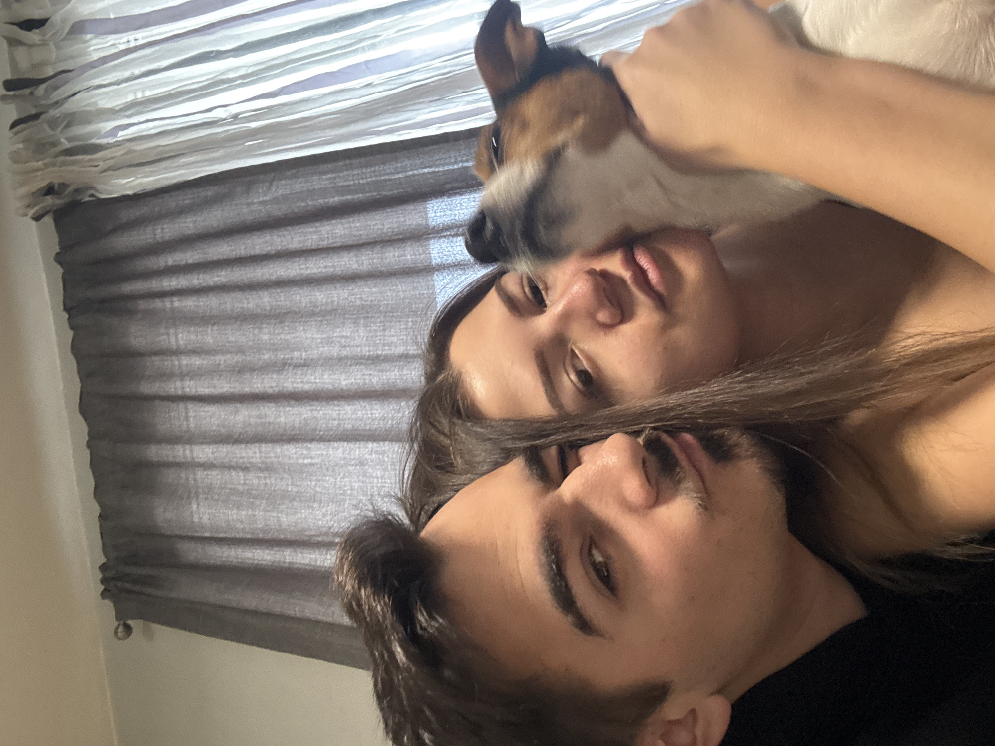 A medium shot of a man and woman lying next to each other with a dog. The woman is lying on her back, and the man is lying slightly below her and closer to the camera. The dog is lying on the woman's chest, with its head near her face. The dog is white and brown. The woman has long brown hair. The man has short brown hair and a beard. There are window blinds in the background. There are no indications of the location being in Sofia, Bulgaria.