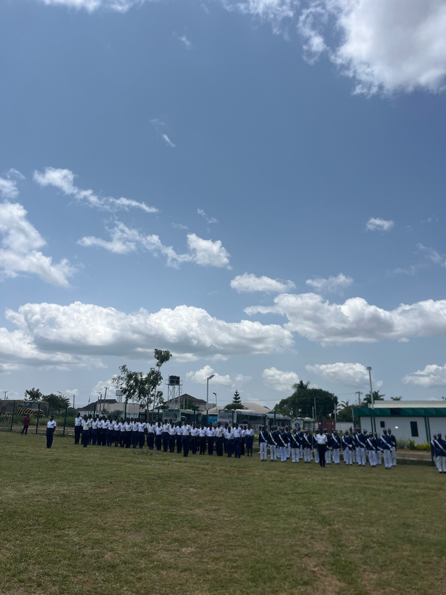 In Barikin Jirga Summa, Nigeria, under a bright, partly cloudy sky, a group of individuals are gathered on a grassy field. A formation of approximately 60 people, dressed in matching dark blue jackets with white trim and white pants, stand in neat rows facing forward. They appear to be participating in an organized event, possibly a parade or ceremony. To the left of the main group, a smaller group of individuals, also in uniform, stand. In the background, several buildings, including one with a green roof, are visible, along with trees and what appears to be a water tower. The scene is set during daylight hours.