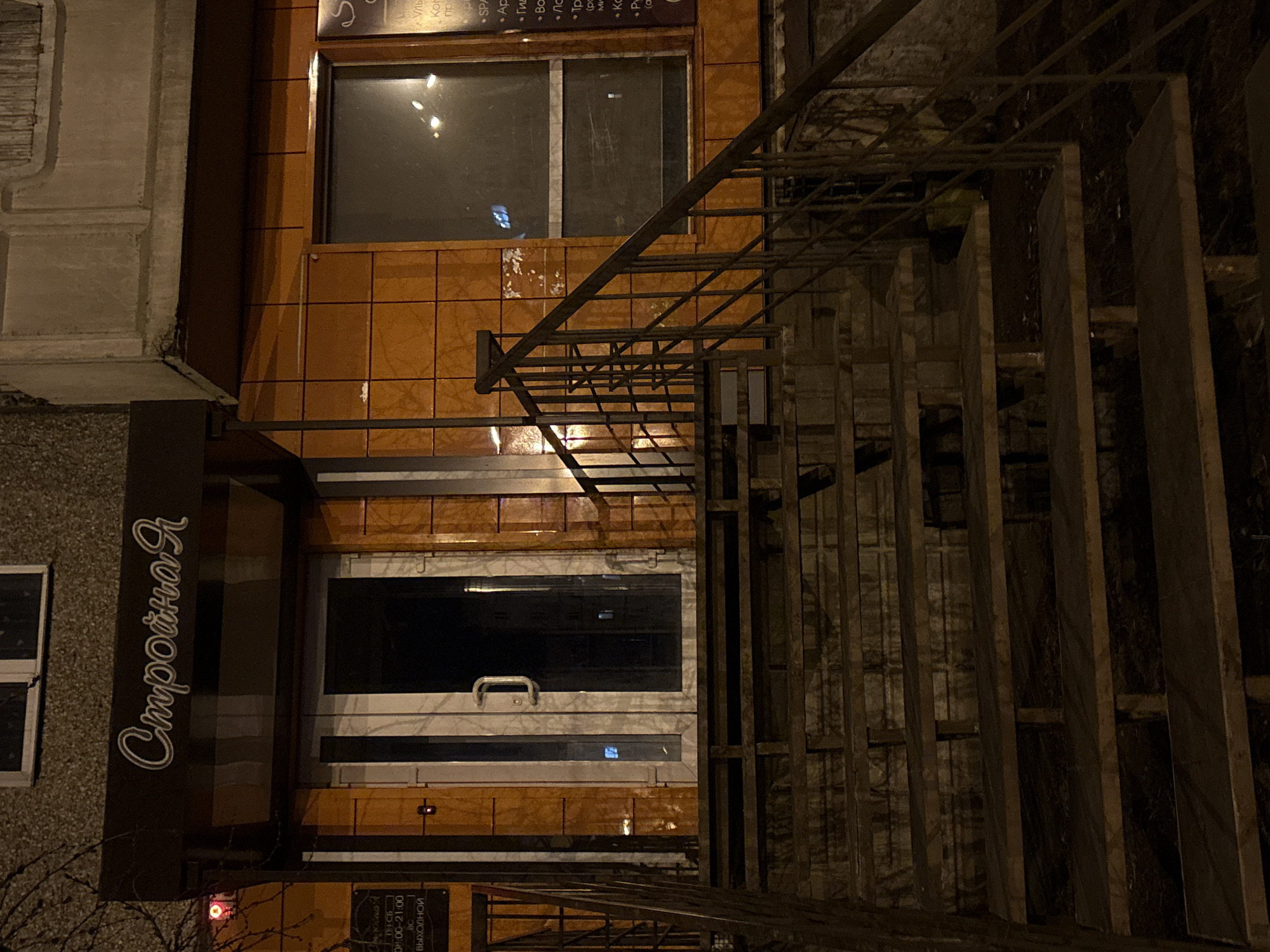 This image depicts the exterior of a building with a sign in Russian that reads "Стройная," which translates to "Slender" or "Slim." The building is constructed with a mix of orange and white panels. The windows appear dark, suggesting it is evening or night. There's a metal staircase or scaffolding structure obstructing the full view of the building, adding depth to the photograph.
There are no people visible in the image. The scene is located in the city of Krasnoyarsk, Russia, as determined by the Russian text on the sign. The OCR of the image also includes the timings for the building which are "ПН-СБ" (Monday-Saturday) 08:00-21:00 (8:00 AM to 9:00 PM) and "BC" (Sunday) "ВЫХОДНОЙ" (closed).