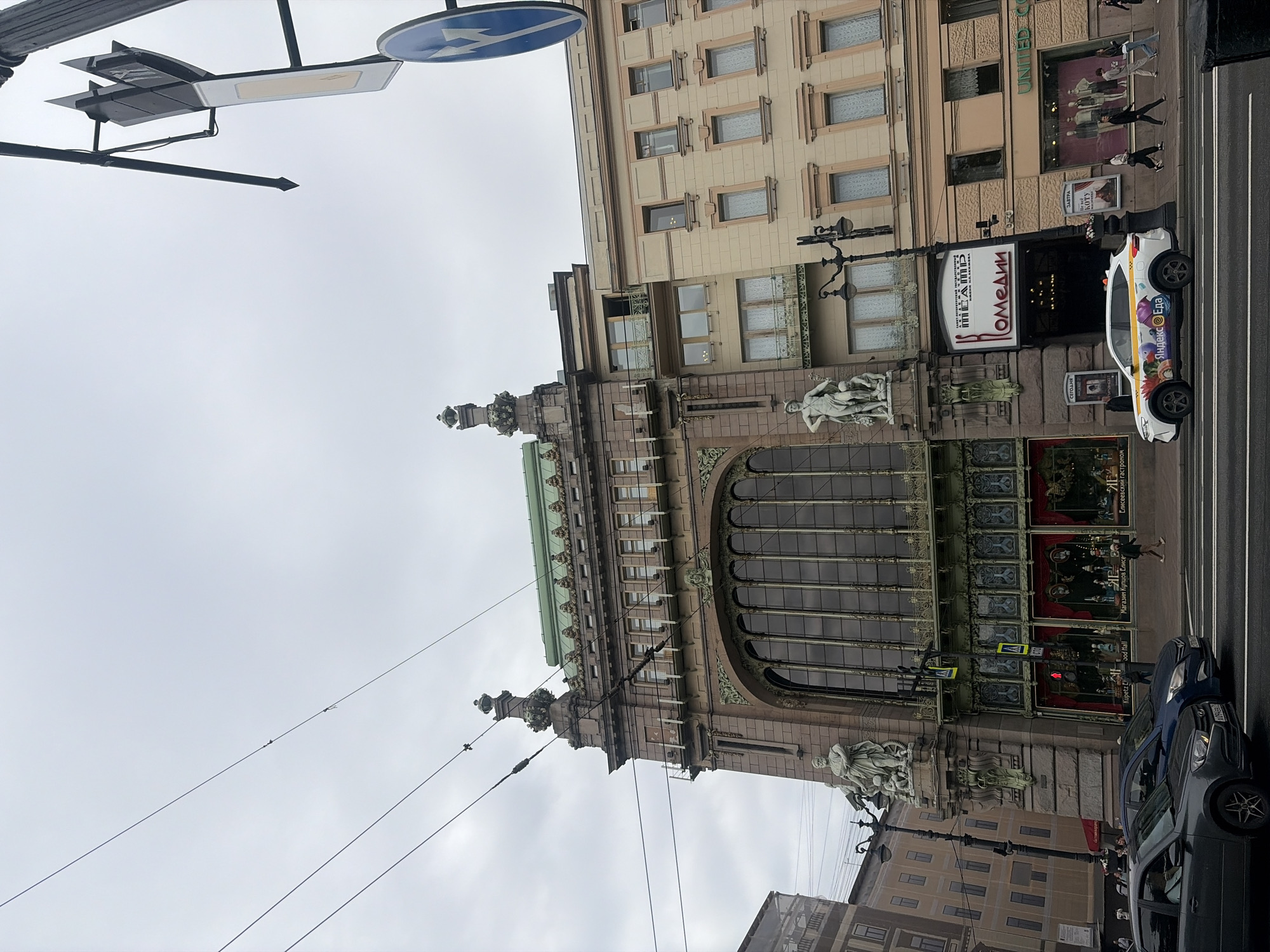 The image shows the Singer House, also known as the Dom Knigi, located on Nevsky Prospekt in Saint Petersburg, Russia. It is a grand, historic building with ornate architecture, featuring a distinctive glass dome and detailed facade. The building is multi-story, with large windows and decorative elements.
In the foreground, a street scene is depicted. Several cars are driving on the wet road, indicating recent rain or a damp environment. A white taxi with colorful advertising is parked near the entrance of a building. Pedestrians are walking on the sidewalk, some entering or exiting establishments.
The sky is overcast, suggesting a cloudy day. Utility wires are visible stretching across the sky, adding to the urban atmosphere. Traffic signs and streetlights are also present, contributing to the functional elements of the city environment. The overall scene captures a typical day in the bustling city of Saint Petersburg, showcasing its architectural heritage alongside modern urban life.