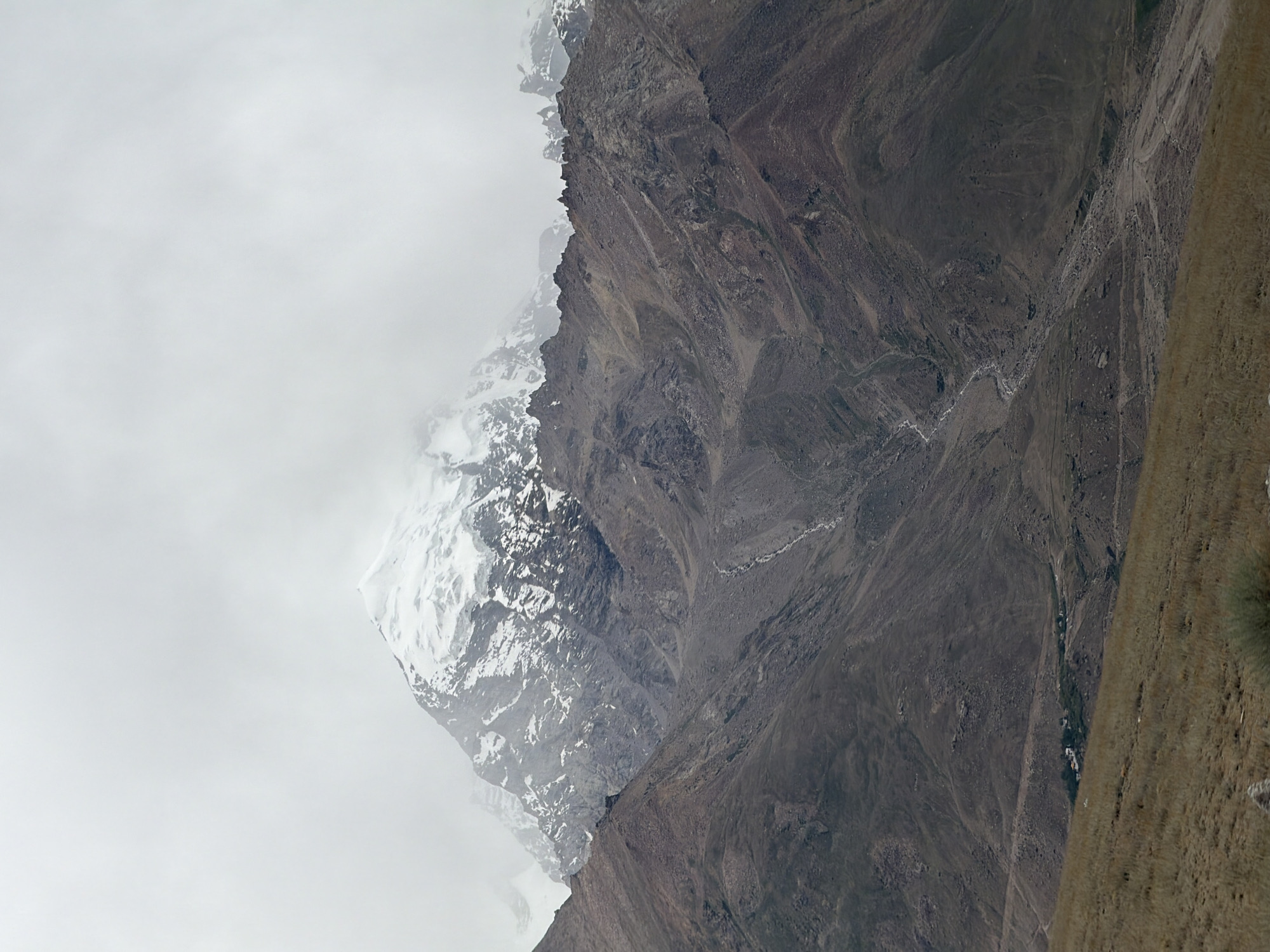 A majestic snow-capped mountain peak pierces through thick, hazy clouds. The mountain's lower slopes are rugged and barren, with visible rock slides and scree. A thin, winding stream or dry riverbed snakes its way down the mountainside. In the middle ground, a small cluster of buildings, likely a village, is nestled against the base of the mountain, surrounded by a patch of green vegetation. The foreground consists of a gently sloping, dry, grassy hillside. The overall scene conveys a sense of remote, high-altitude wilderness. The image depicts the landscape near Pibiting, India.