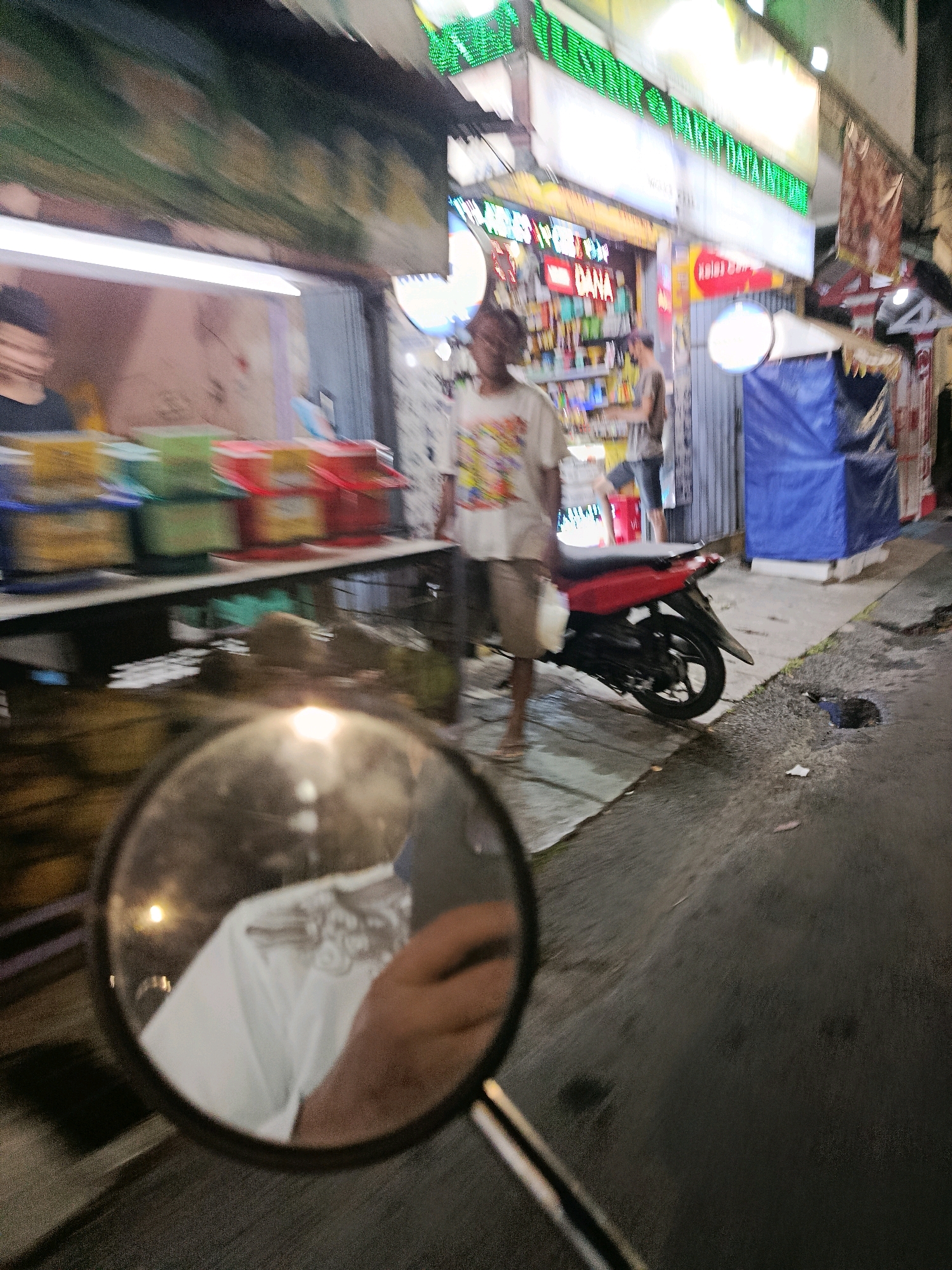 The image depicts a street scene at night, possibly in Bekasi, Indonesia, characterized by a food stall on the left and a sidewalk with a red motorcycle parked on the right. In the foreground, a rearview mirror reflects a person wearing a white t-shirt and holding a white plastic bag. The food stall displays colorful plastic containers filled with food items, and a fluorescent light illuminates the area. Two individuals are visible near the stall; one appears to be a vendor, and another is standing by the motorcycle. The street is wet, suggesting recent rain or a cleaning operation. Several large, round objects, possibly coconuts, are also present near the stall. The overall atmosphere is dimly lit, with artificial lights from the stall and overhead signs creating highlights and shadows.