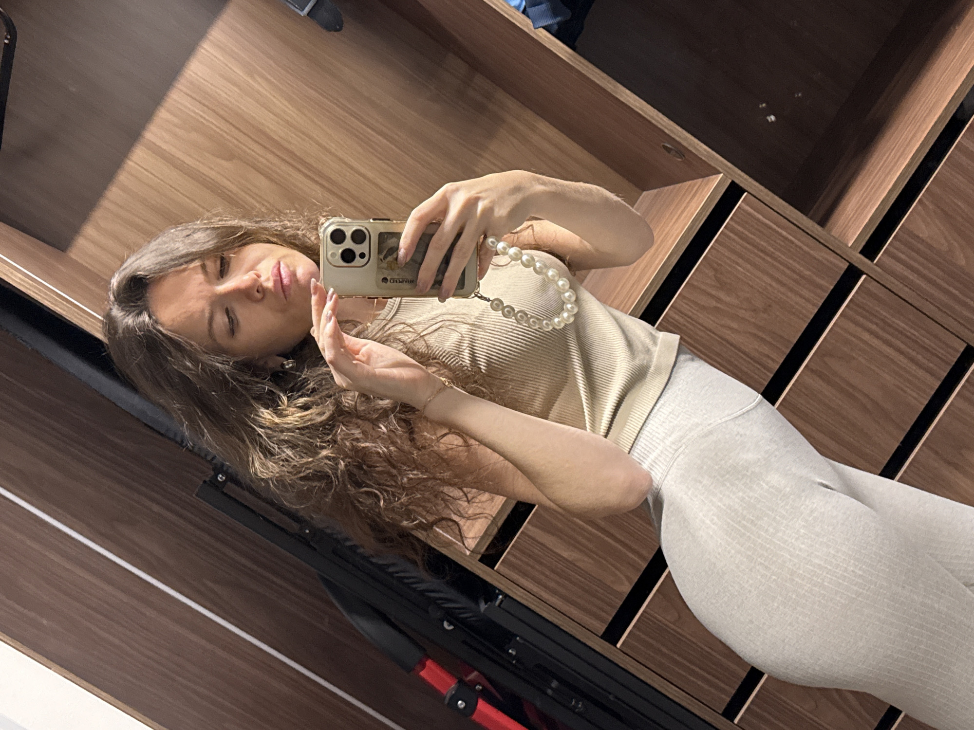 A mirror selfie captures a young woman indoors, likely in Moscow, Russia. She has long, wavy light brown hair and is dressed in a light beige ribbed top and light grey ribbed leggings. Her left hand holds a light-colored smartphone, possibly an iPhone, adorned with a pearl strap accessory and a case featuring the text "BELIEVЯ" and a classical painting fragment. She is making a "duck face" expression with her eyes partially closed, looking towards the camera.
The background is dominated by warm-toned wooden cabinetry or a wardrobe with distinct black vertical and horizontal lines. A prominent diagonal shadow falls across the upper left portion of the wooden surface, indicating a light source. In the lower left, a dark object with red accents is partially visible, possibly part of furniture or equipment. The overall scene is brightly lit, emphasizing the textures of her attire and the wooden interior.