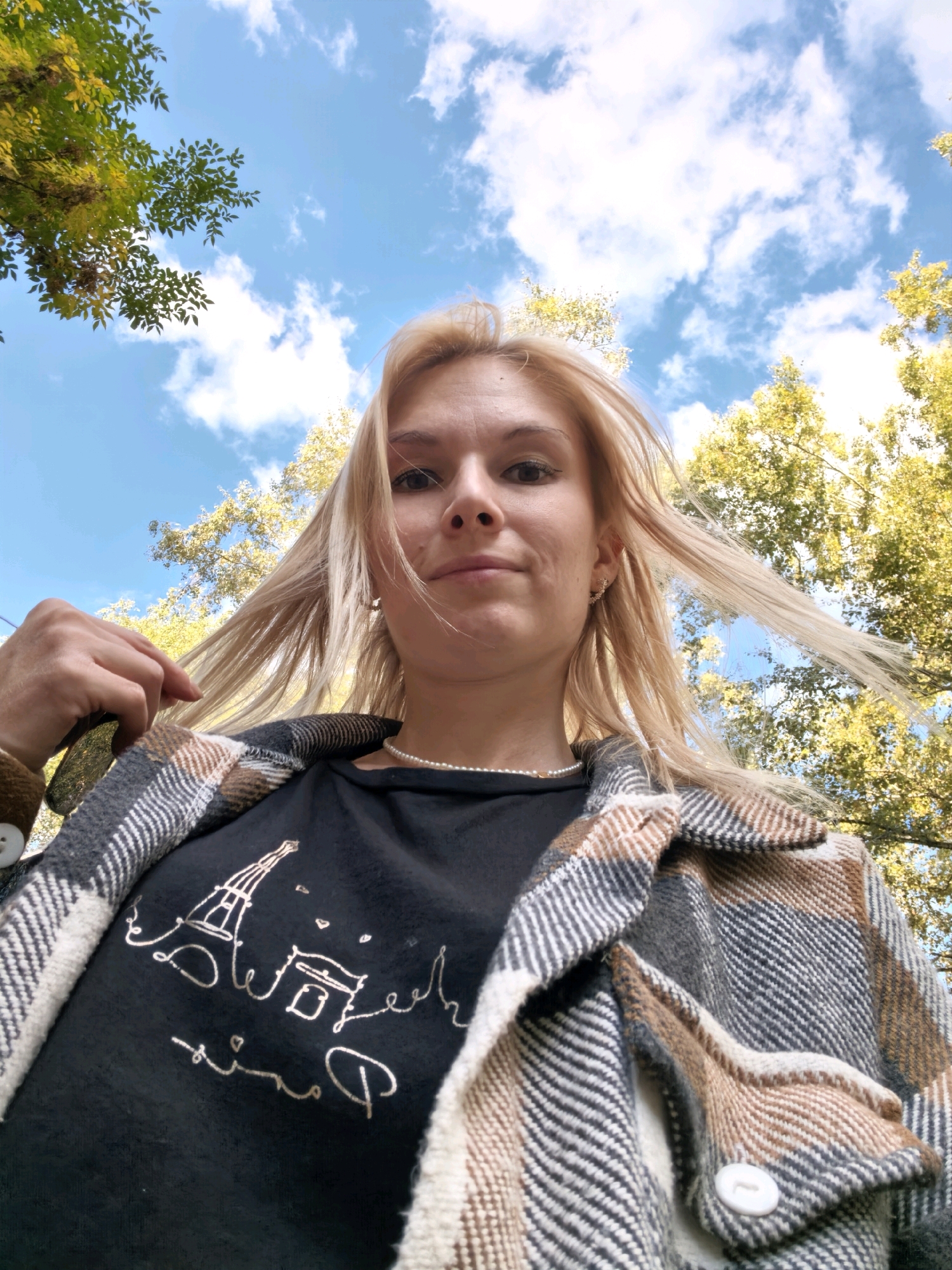 A young woman with blonde hair and fair skin is captured in a low-angle shot, looking upwards towards a blue sky with scattered clouds and surrounded by the yellowing leaves of autumn trees. She is wearing a black t-shirt with a silver outline drawing of the Eiffel Tower and Parisian-themed text, and a plaid jacket with brown, grey, and white tones. Her left hand is raised, gently pulling at her hair, and she is wearing a pearl necklace and small earrings. The background suggests an outdoor setting, possibly a park or a tree-lined street. There is no discernible evidence to place this image specifically in the city of Sterlitamak, Russia.