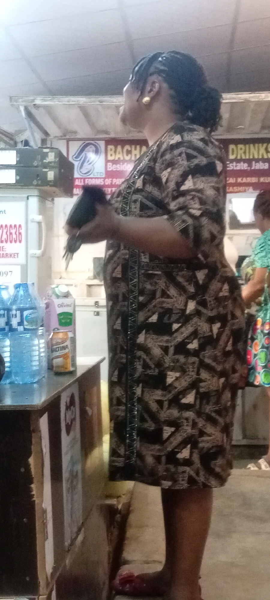 A medium shot shows a woman standing in what appears to be a small store. She is wearing a patterned brown and beige dress, gold earrings, and her hair is styled in braids pulled back into a bun. In her hands, she holds a dark object.
Behind her, signs advertise "BACHA" with the text "Beside ALL FORMS OF PAY" and "DRINKS Estate, Jaba." A refrigerator is visible to the left with a phone number beginning with "23636." Water bottles, Chivita juice, and Maltina soda cans sit on a wooden counter in the foreground.
The woman stands barefoot in pink sandals with visible bare legs. Another woman, partially visible in a green shirt and patterned skirt, is standing in the background.
Given the limited information from the image, I cannot confirm the location in the city of Kano, Nigeria.