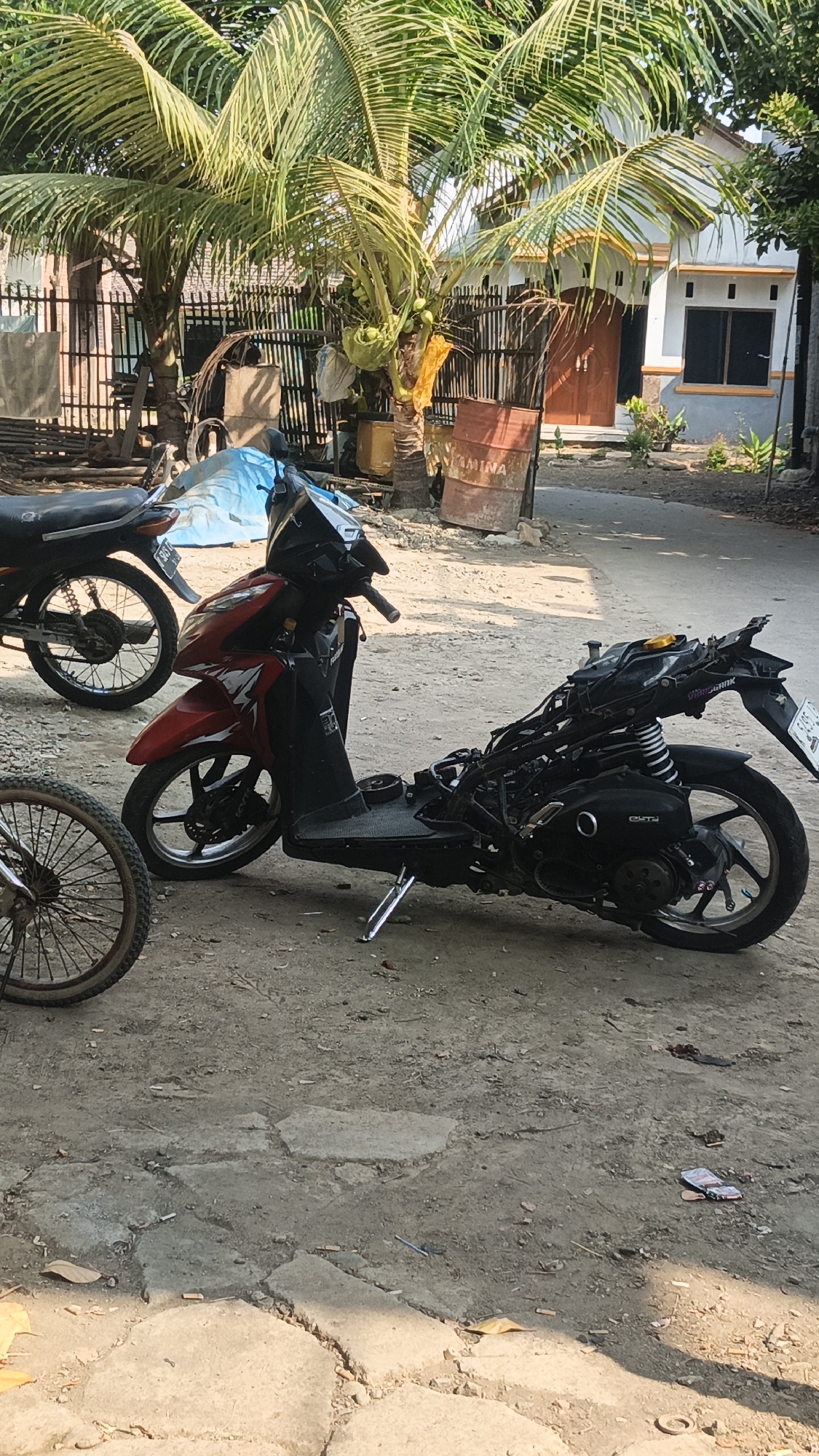The image depicts a street scene in Bulungcangkring, Indonesia. In the foreground, a red and black scooter is partially disassembled, with its engine and rear section exposed. To the left of the scooter, a bicycle wheel with a worn tire is visible. Behind the scooter, another motorcycle, mostly obscured by the red scooter, is parked. The background features a residential dwelling with a light-colored facade and a dark wooden door, partially shaded by mature palm trees with coconuts. A bamboo fence runs along the property line, and various objects, including a blue tarp and a rusty barrel, are present in the yard. The ground is a mixture of dirt and cracked concrete, with scattered debris, including cigarette butts and fallen leaves. Sunlight casts shadows across the scene, indicating it is daytime.