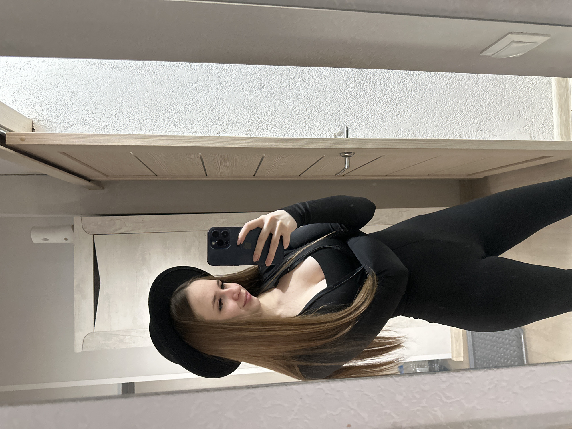 A woman in a black jumpsuit and a black hat is taking a mirror selfie. Her long, light brown hair is partially covering her face. She is holding a dark-colored smartphone in her left hand, which is positioned slightly above her waist. The background includes a white wall and a light-colored wooden cabinet with metallic hardware. The location is Unknown, Unknown.