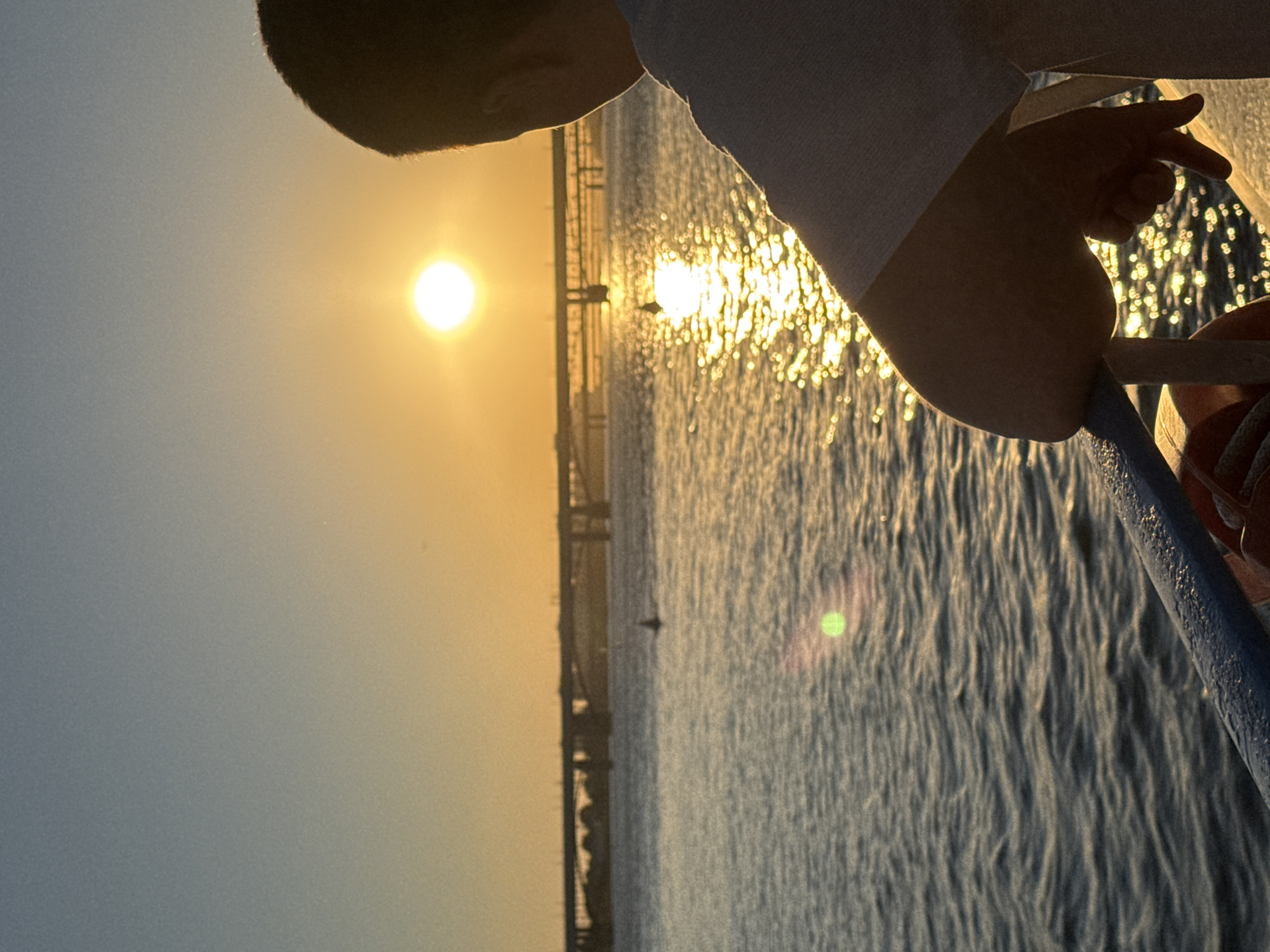 A lone figure, identified as a young male, stands on a vessel, gazing towards a sunset over a body of water. The sun, a brilliant orb, is low on the horizon, casting a warm golden glow across the scene and creating a shimmering reflection on the water's surface. In the distance, a bridge spans the water, hinting at the urban landscape of Novosibirsk, Russia. The atmosphere is serene and contemplative, with the gentle ripples of the water and the fading light contributing to a tranquil mood. The boy's posture suggests a moment of quiet reflection as he observes the natural beauty unfolding before him.