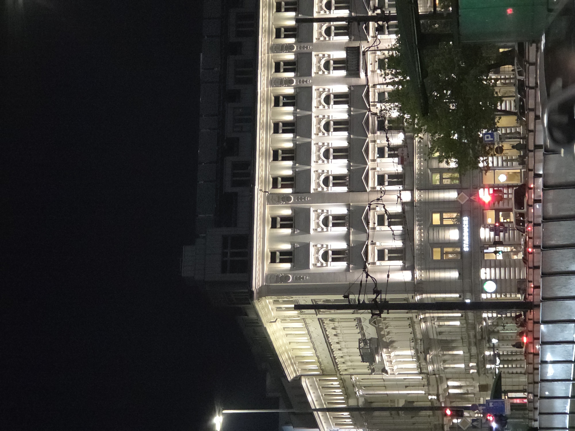 The image shows a night scene in Warsaw, Poland. A brightly lit, ornately designed building with multiple windows and detailed architectural features is visible. A Starbucks logo is prominently displayed on one section of the building. Streetlights and traffic signals are present, including red lights indicating a stop. Wires and poles suggest the presence of an overhead electrical or telecommunications infrastructure. The left side of the image is mostly dark, showing the outline of another building with a simpler design in the background. A tree is also visible, partially obscuring some windows of the ornate building.