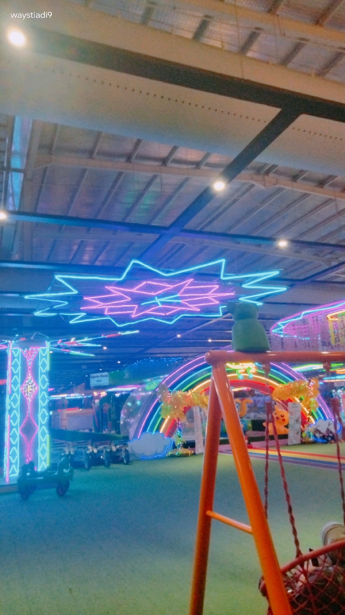 The image depicts an indoor playground or entertainment center, likely in Bandung City, Indonesia, given the watermark "waystiadi9." The scene is characterized by vibrant neon lighting, primarily in blue and pink, outlining star shapes on the ceiling and various attractions. In the foreground, an orange swing set with a red net seat is visible, with the right side of the frame occupied by the legs of a person seated on the swing. Behind the swing, a large, illuminated rainbow archway is prominent, with decorative elements such as yellow foliage and cartoonish clouds. Further back, other attractions and structures are visible, including a set of toy vehicles and tall, illuminated decorative pillars. The floor appears to be covered in artificial green turf, typical of indoor play areas. The overall atmosphere is playful and colorful, illuminated by artificial light.