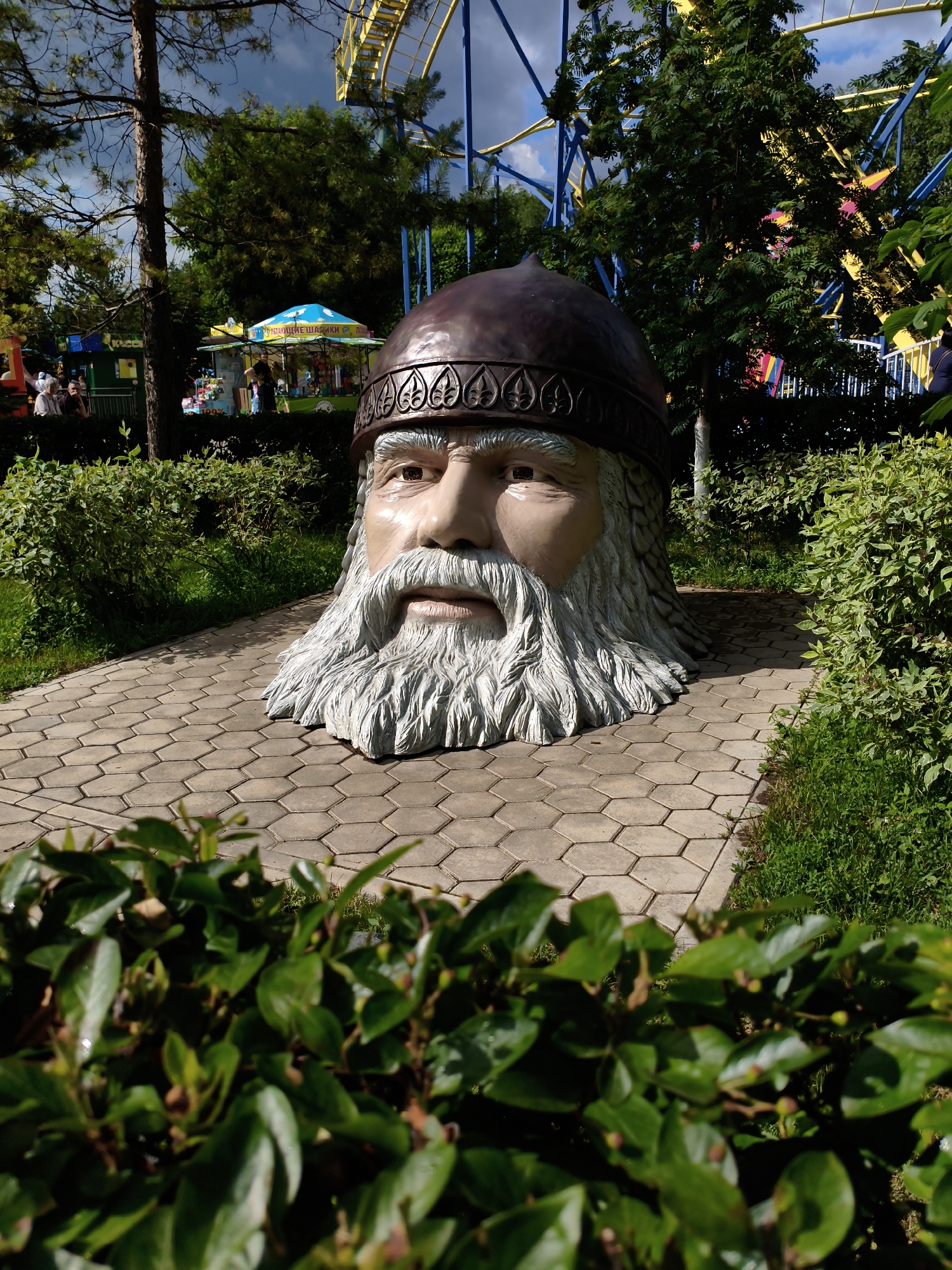 In Orenburg, Russia, a large, weathered sculpture of a man's head with a distinguished beard and helmet is positioned on a hexagonal paved pathway. The sculpture is surrounded by lush green bushes and trees, with glimpses of an amusement park in the background. A vibrant yellow roller coaster track is visible overhead, suggesting the sculpture is located within or near an amusement park or entertainment venue. The sky is partly cloudy, indicating a pleasant day. There are no people directly interacting with the sculpture, though distant figures can be seen in the amusement park area. The overall scene depicts an outdoor recreational space with a prominent artistic element.
