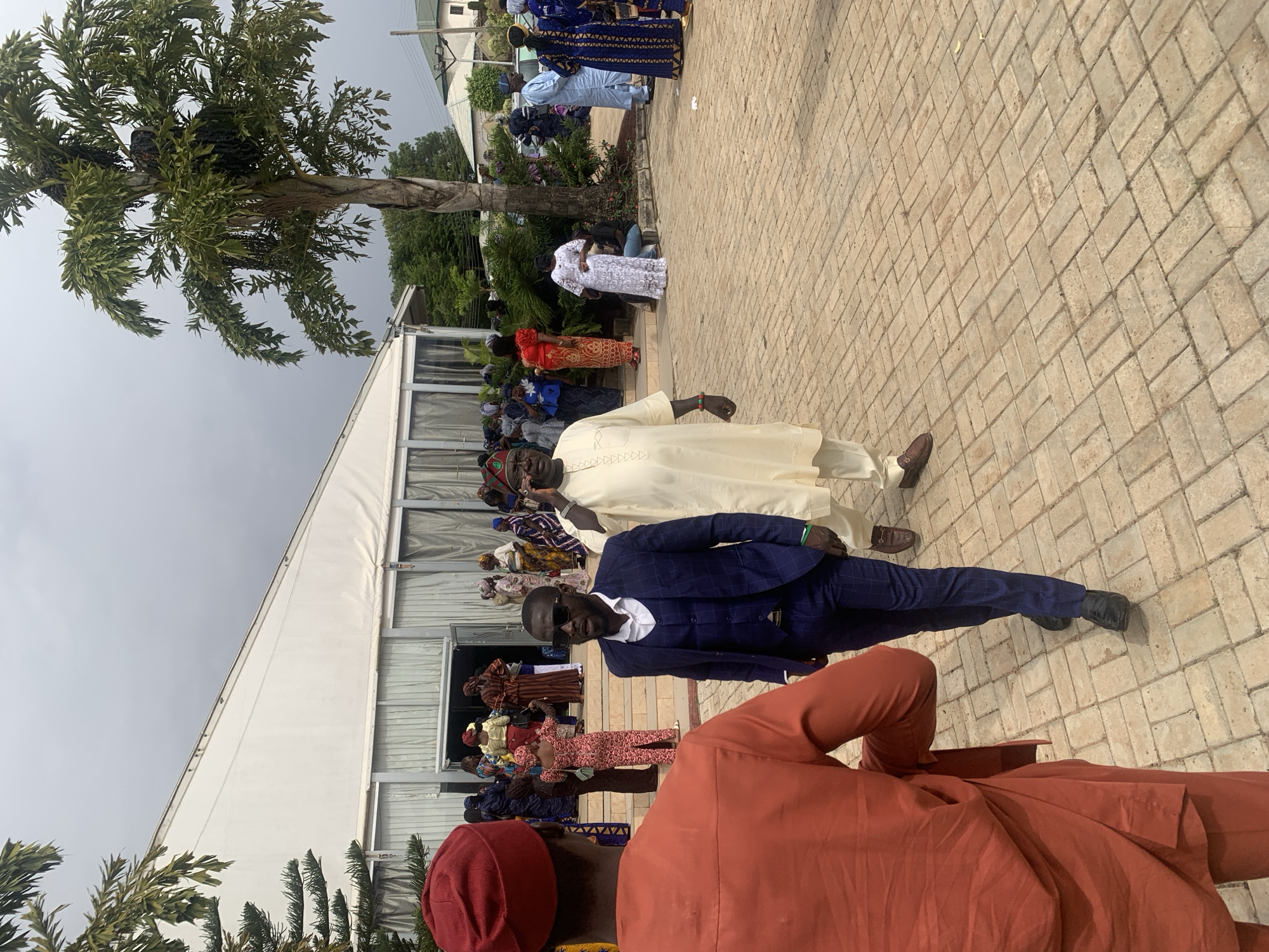 The image shows a gathering of people outside a building in Jos, Nigeria. The architecture suggests a modern event space. A palm tree stands to the left of the building. The ground is paved with light brown brick.
Several individuals are visible, including men dressed in both traditional attire (cream-colored flowing garments, red hats) and modern suits. The people are standing or walking. Other people are congregated near the building's entrance. The sky is overcast.