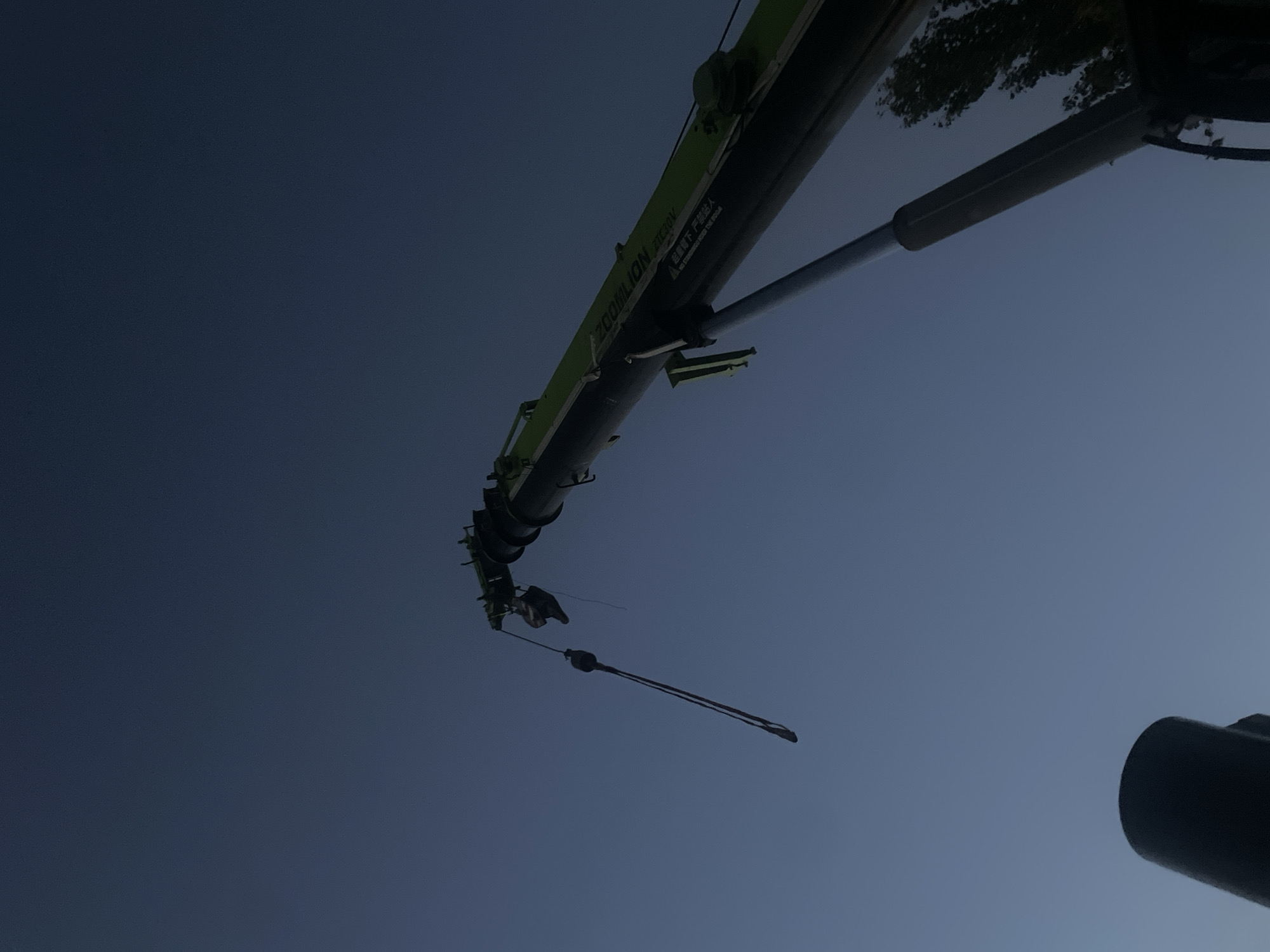 A low-angle shot captures a large green crane against a dusky blue sky. The crane's boom extends upwards and to the right, with a hook and cable hanging down from its end. Text is visible on the crane's boom, including "ZOOMILION ZTC300V". A hydraulic cylinder is also visible extending from the base of the crane's boom. In the lower left corner of the image, a portion of another dark, cylindrical object, possibly part of the crane's base or another piece of equipment, is visible. A hint of a tree with dark foliage is present on the far right of the image, suggesting an outdoor environment. The lighting indicates it is either dawn or dusk. The scene is not identifiable as being specifically within Bishkek City, Kyrgyzstan without further contextual information.