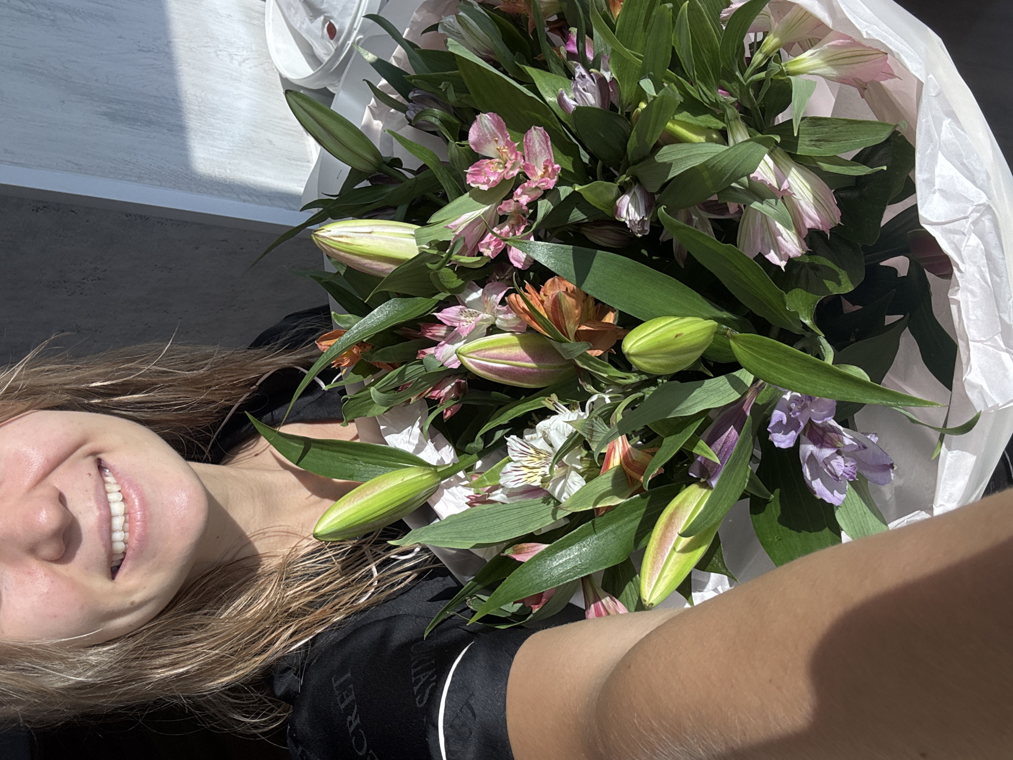 A selfie shows a fair-skinned person smiling and holding a bouquet of lilies in their lap. The bouquet is wrapped in white paper and contains various colors of lilies, including pink, orange, purple, and white, as well as green leaves and unopened buds. The person is wearing a black garment with white lettering visible. The background includes a window with sunlight streaming in, casting shadows on a light gray surface. The location is unknown.