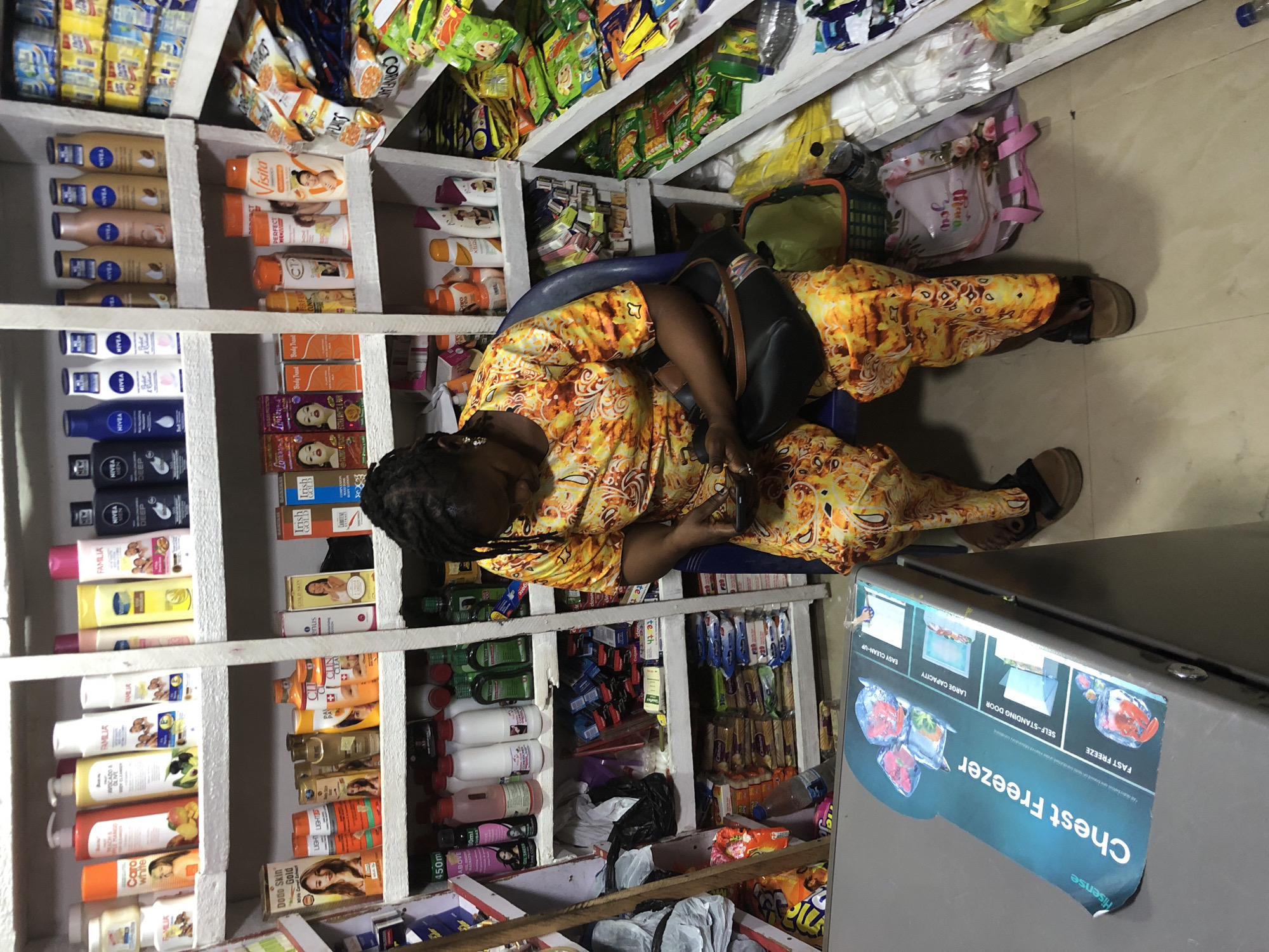 A Nigerian woman in an orange and yellow patterned dress sits on a blue plastic chair in a small convenience store. She is looking down at her phone, which she holds in her right hand. In front of her is a gray chest freezer with a Hisense "Chest Freezer" label. The store shelves are stocked with various products, including soaps, lotions, drinks, and snacks. The lighting is bright and artificial. The floor is tiled.