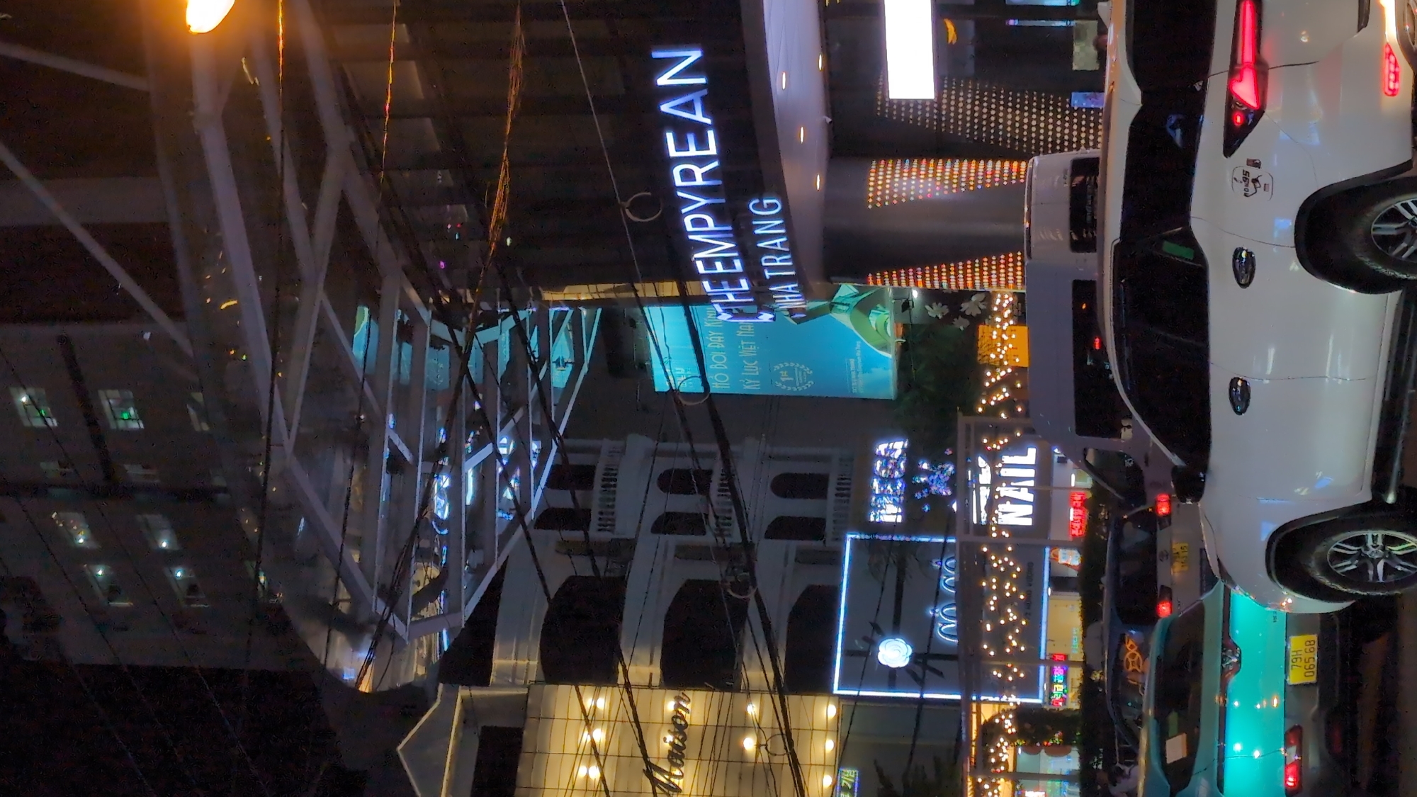 The image shows a nighttime street scene in Nha Trang, Vietnam. A white SUV is prominently featured in the foreground, parked on a street. In the background, a building with the illuminated signage "THE EMPYREAN NHA TRANG" is visible. Other buildings with glowing signs and lights are also present, along with overhead power lines and a pedestrian walkway structure. The scene appears to be a busy urban environment with vehicle and building lights illuminating the night.
