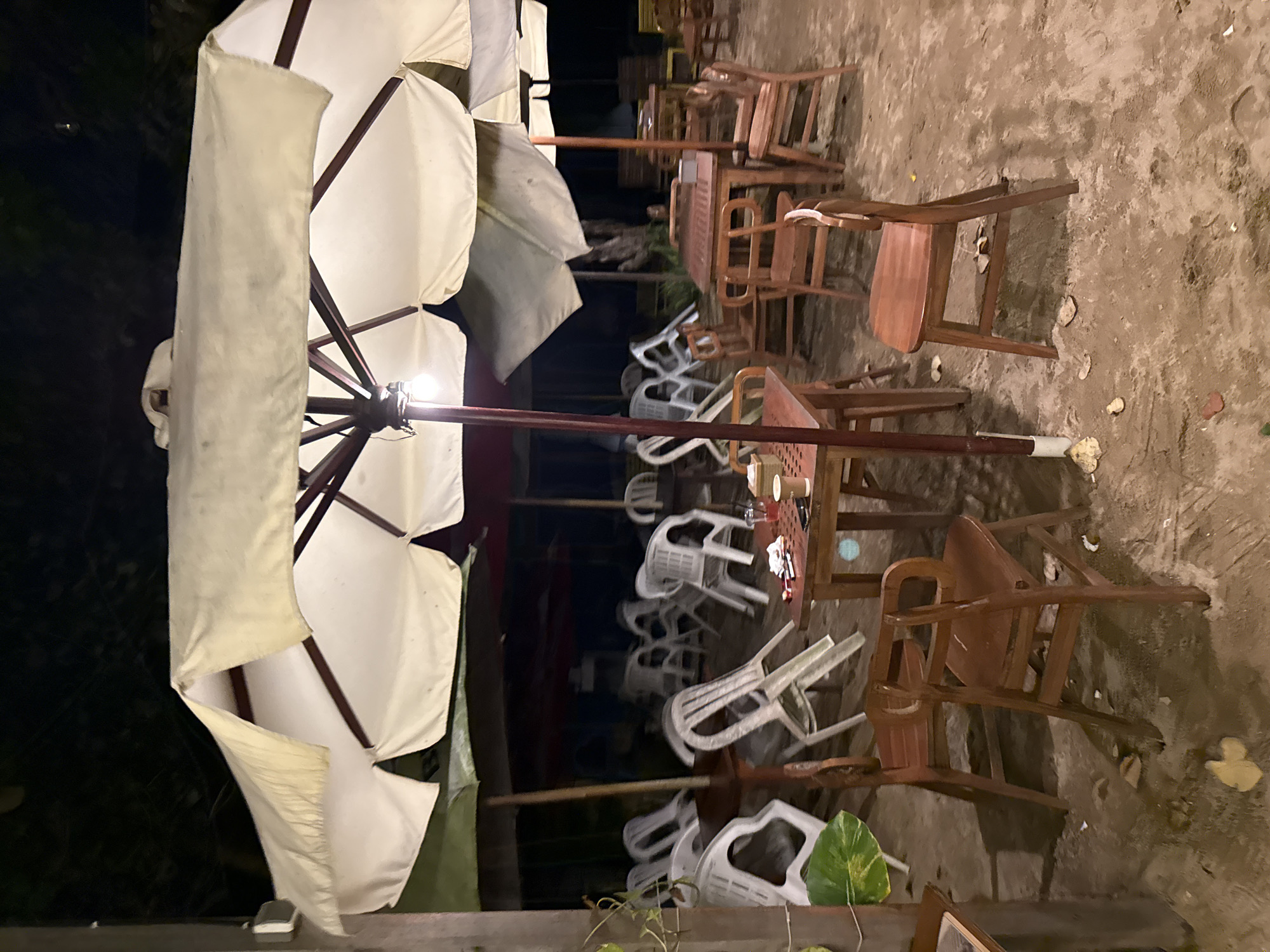 A night-time outdoor scene depicts an empty beachside cafe in Denpasar, Indonesia. Multiple tables and chairs, some made of wood and others of plastic, are scattered across a sandy ground. Large umbrellas, some white and some green, provide shade. The lighting appears to be artificial, with a visible light bulb under one of the umbrellas. There are no people present. A few drinks and some napkins are visible on one of the tables. The environment suggests a closed establishment, possibly after closing hours or during a quiet period.