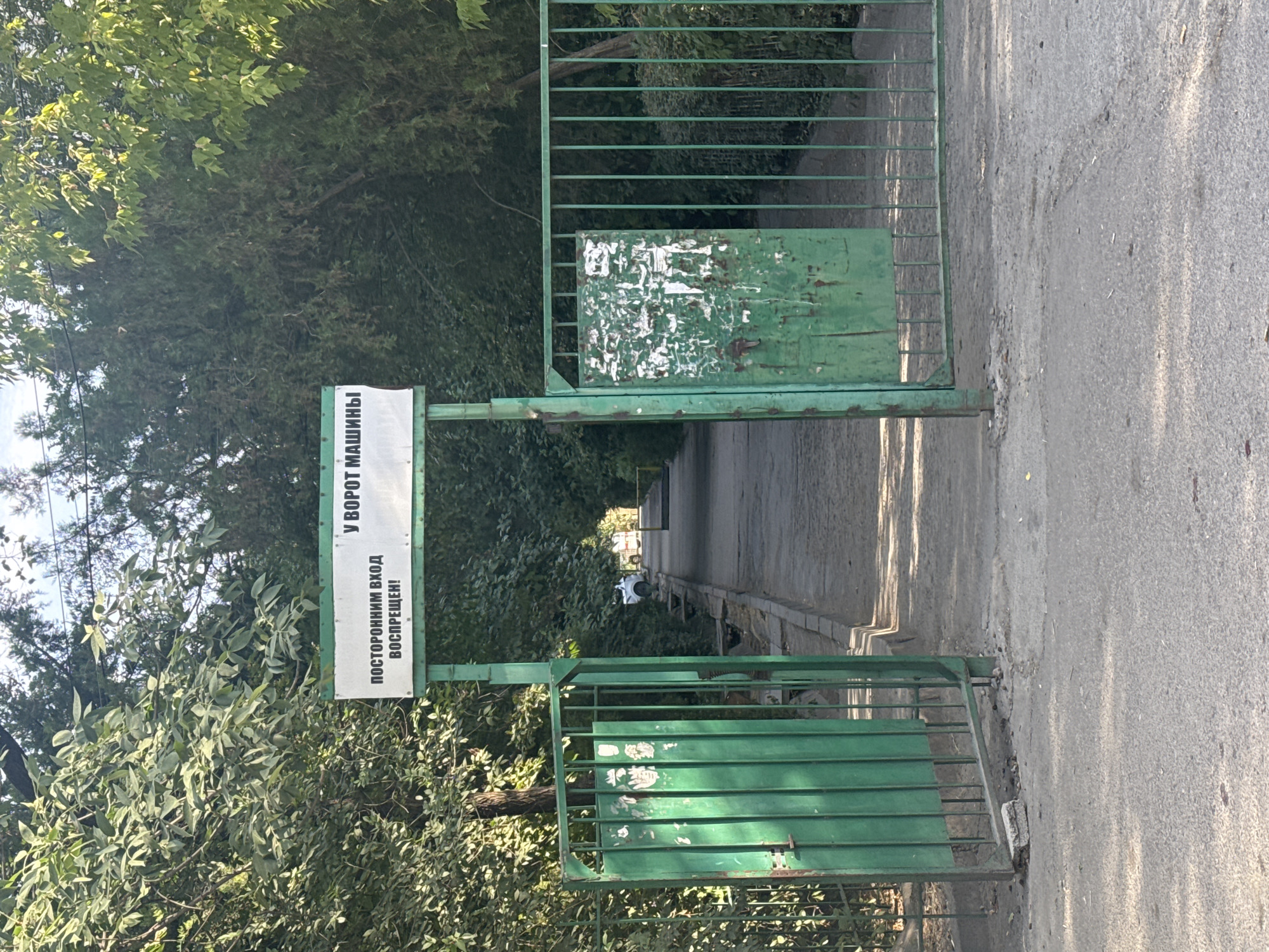 An open green metal gate stands at the entrance to a tree-lined pathway. Above the gate, a sign reads "В У ВОРОТ МАШИНЫ" and below it, "ПОСТОРОННИМ ВХОД ВОСПРЕЩЕН!" (English translation: "CARS AT THE GATE" and "STRANGERS ENTRY FORBIDDEN!"). The pathway extends into the distance, with trees on either side. A white car is visible parked on the side of the pathway. The scene appears to be in a park or a residential area. The overall impression is one of a controlled or private entrance.