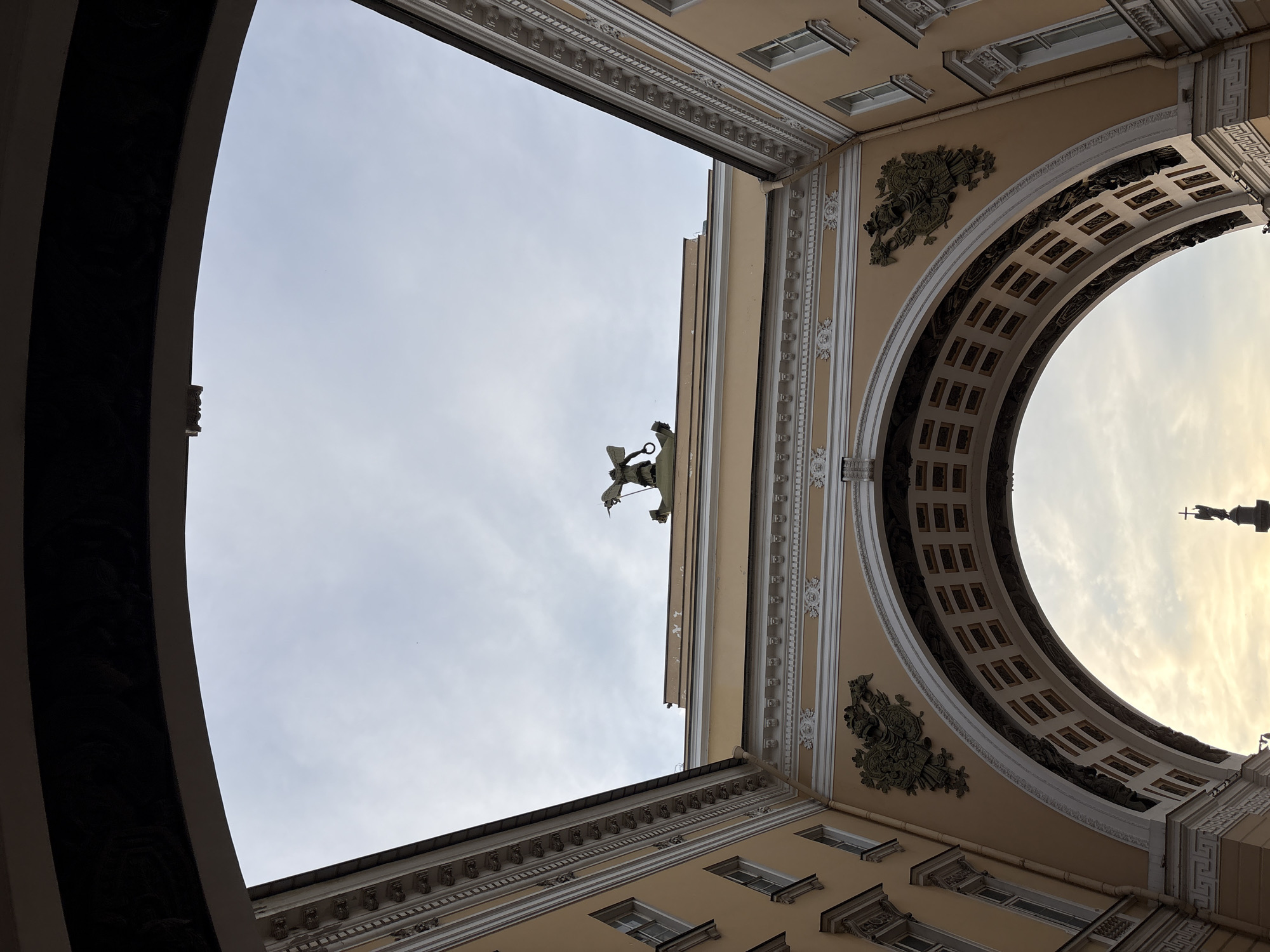 The image depicts a low-angle view of the Triumphal Arch of the General Staff Building in Palace Square, Saint Petersburg, Russia. The arch, constructed from a pale yellow stone, is grand and ornate, featuring decorative carvings and a coffered interior. Atop the arch, a sculptural group depicts an angel with wings outstretched, holding a staff. The sky above is overcast with a hint of sunset light breaking through the clouds. The architectural details of the surrounding buildings, with their windows and decorative cornices, are visible. In the distance, through the arch, a faint silhouette of a tall column with a statue is discernible, likely Alexander Column. The scene is devoid of people.