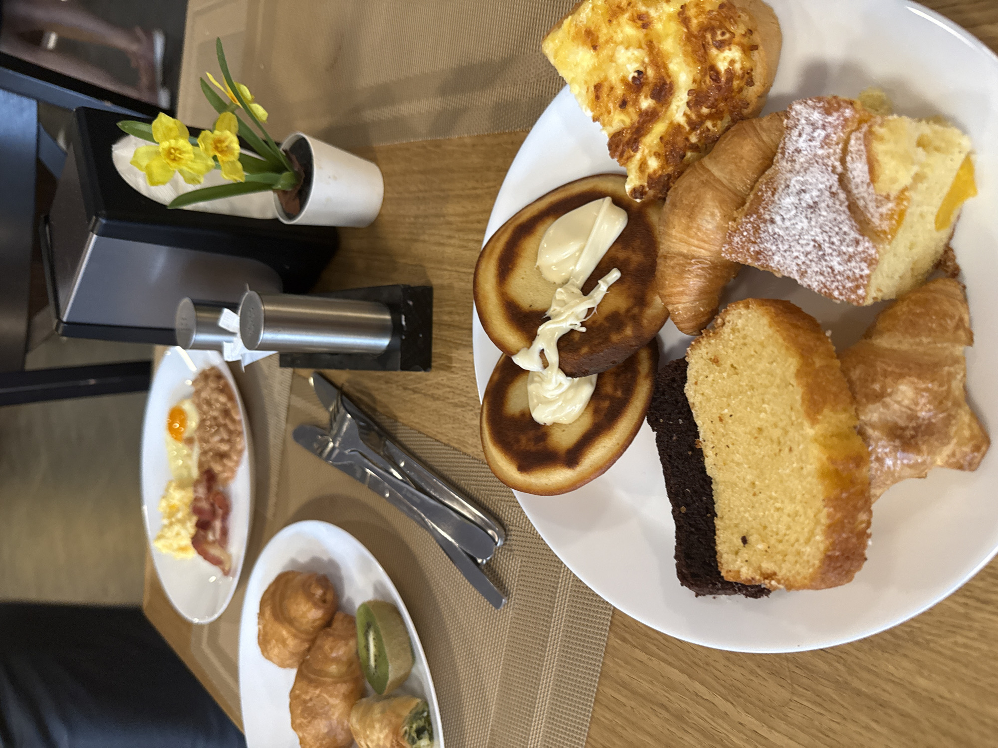 The image shows a table setting with several plates of food, likely in a hotel or restaurant, location unknown. A large white plate in the upper right contains pancakes with white cream, a croissant, a pastry dusted with powdered sugar, a yellow cake slice with a dark layer, an omelet, and another croissant. To the left, a smaller white plate holds scrambled eggs, bacon, and fried eggs. Below that, a third white plate has croissants and slices of kiwi. Between the plates are cutlery sets. Adjacent to the first plate, there is a black tray with a salt and pepper shaker, and a small white vase holding yellow daffodils. The tabletop has a light brown, textured placemat.