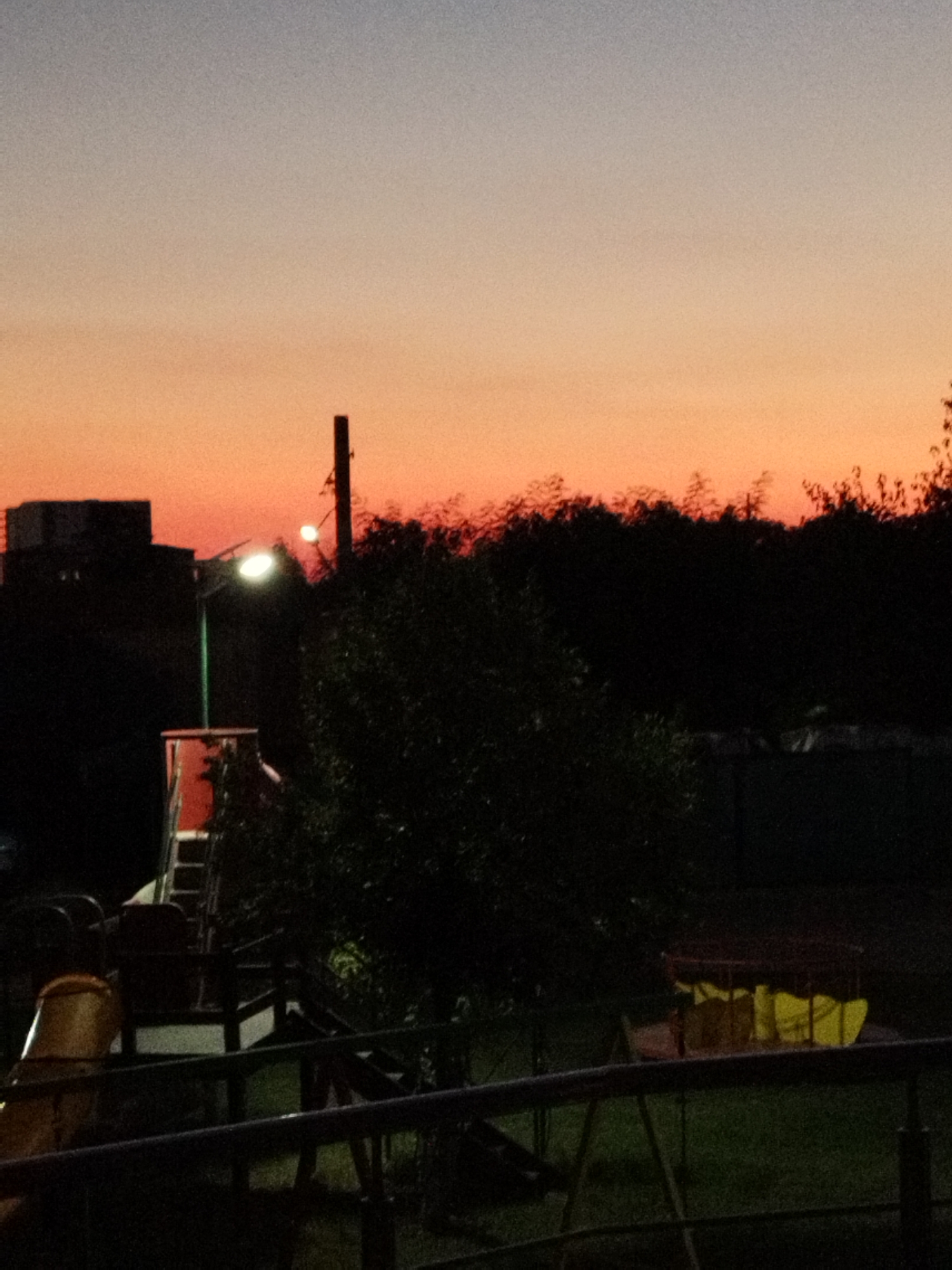 The image depicts a twilight scene with a gradient sky transitioning from a pale blue at the top to vibrant orange and pink near the horizon. A silhouetted treeline runs across the mid-ground, obscuring any details of what lies beyond. In the foreground, a dark silhouette of a playground structure is visible, including what appears to be a slide and some seating. Two streetlights are illuminated, one with a bright white light on a pole and another with a softer glow further in the background. On the left side of the image, a dark building mass with some illuminated windows is also silhouetted against the sky. The overall atmosphere is calm and serene, with the natural light of dusk creating a sense of peacefulness.
