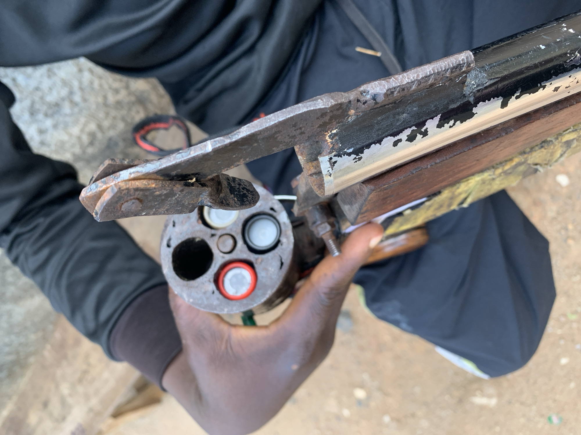 A person is holding a homemade, multi-barreled firearm. The firearm consists of multiple metal tubes arranged in a circular configuration and attached to a stock, with a firing mechanism visible. The ammunition appears to be self-made, likely cartridges with red or white primers. The location is New Stadium, Nigeria, based on provided information. The person is sitting.