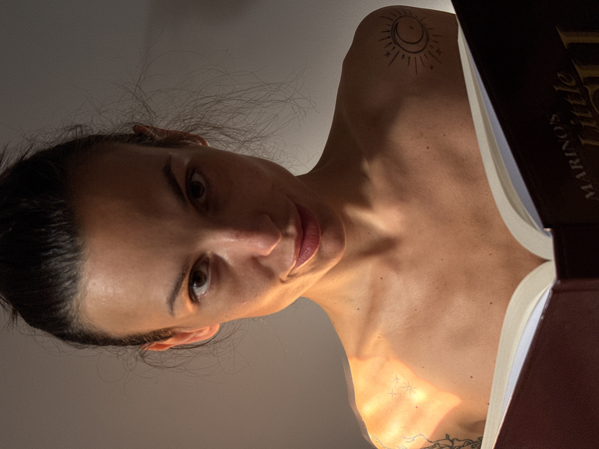 A woman is looking down, holding a book open in front of her. The book's cover is visible, and part of the title seems to read "MARINO'S Little." The woman has her hair pulled back. A tattoo of a crescent moon and sun is visible on her right shoulder. There is another tattoo on her left shoulder. No location information is in the image.