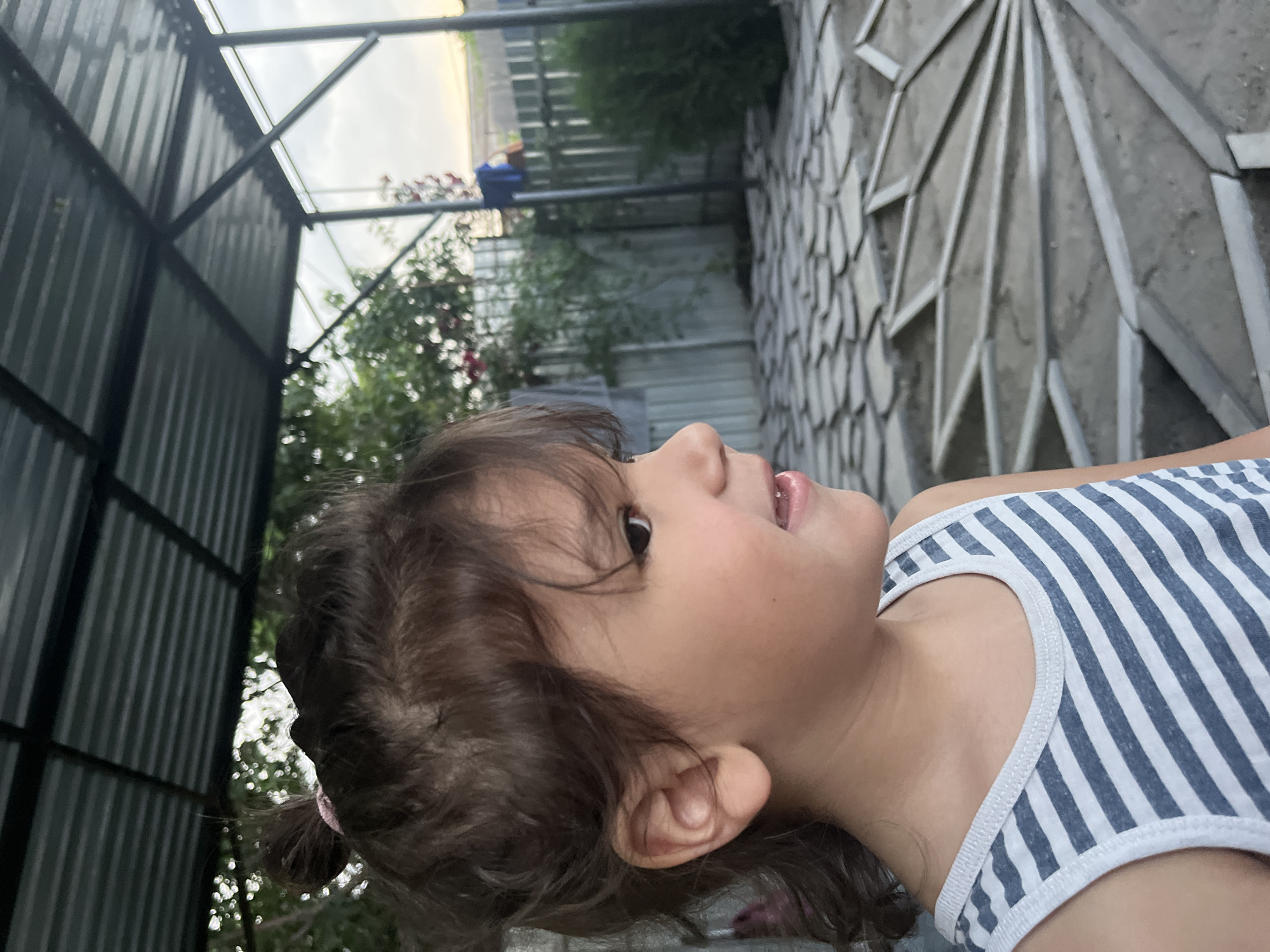 A young girl with brown hair tied in a bun is shown in profile, looking to the right. She is wearing a blue and white striped tank top. The background suggests an outdoor setting, possibly a backyard or patio area, with a corrugated metal roof overhead, a fence, greenery, and a paved area with a geometric pattern. The lighting indicates it is daytime, likely late afternoon or early evening due to the soft light.