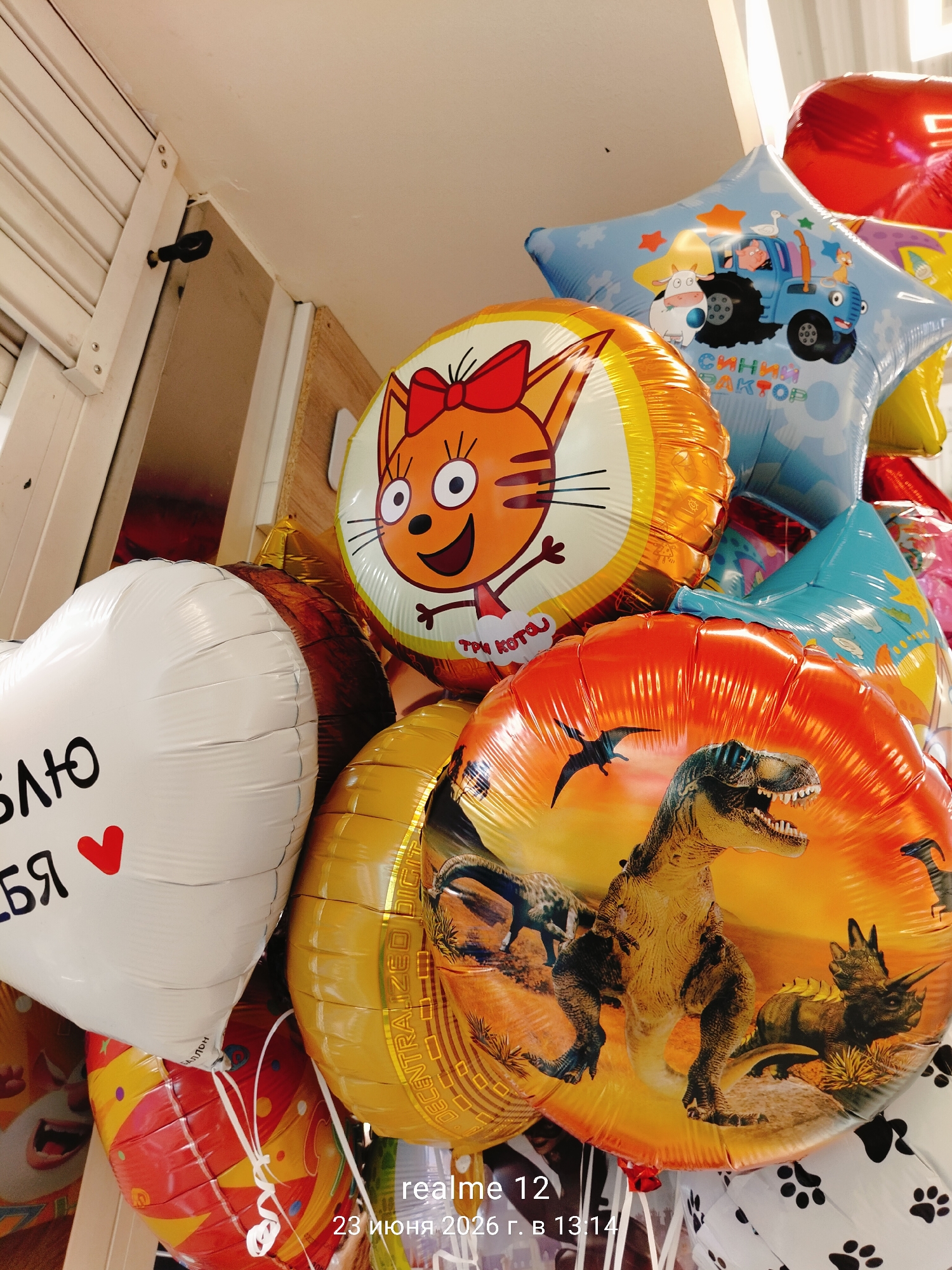 This media file shows a close-up view of a collection of Mylar balloons, likely intended for a celebration. The balloons are a mix of shapes and designs, including a heart-shaped white balloon with black text that partially reads "БЯ лю" and a red heart. Other balloons feature cartoon characters, such as an orange cat with a red bow, a blue star-shaped balloon with a cartoon tractor and animals, and a round balloon with a dinosaur theme, depicting a Tyrannosaurus Rex and other prehistoric creatures. The balloons are clustered together, with some appearing to be attached by white ribbons. The background is slightly blurred but suggests an indoor setting, possibly a store or a room where decorations are being prepared. The lighting appears to be artificial, creating reflections on the shiny surfaces of the balloons.
Visible text on the balloons includes "СИНИЙ ПАКТОР" which translates to "BLUE TRACTOR", and "БАЛЛОН" which means "BALLOON". There is also text partially visible on the white heart balloon. A timestamp in the lower right corner of the image reads "realme 12 23 июня 2026 г. в 13:14", indicating the date and time the photo was taken. The overall impression is one of festive anticipation.